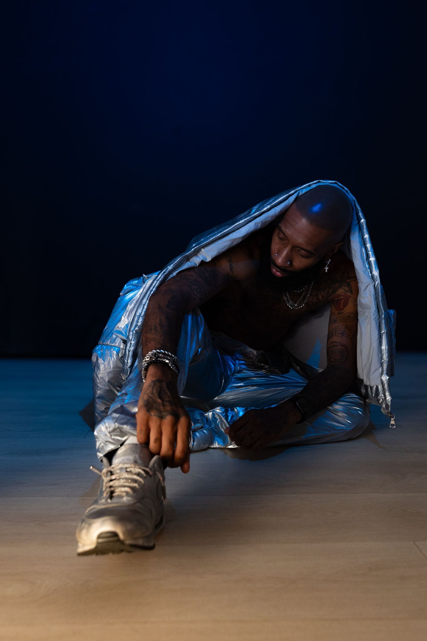 Tattooed man sitting on floor wrapped in silver metallic fabric against a dark blue background.
