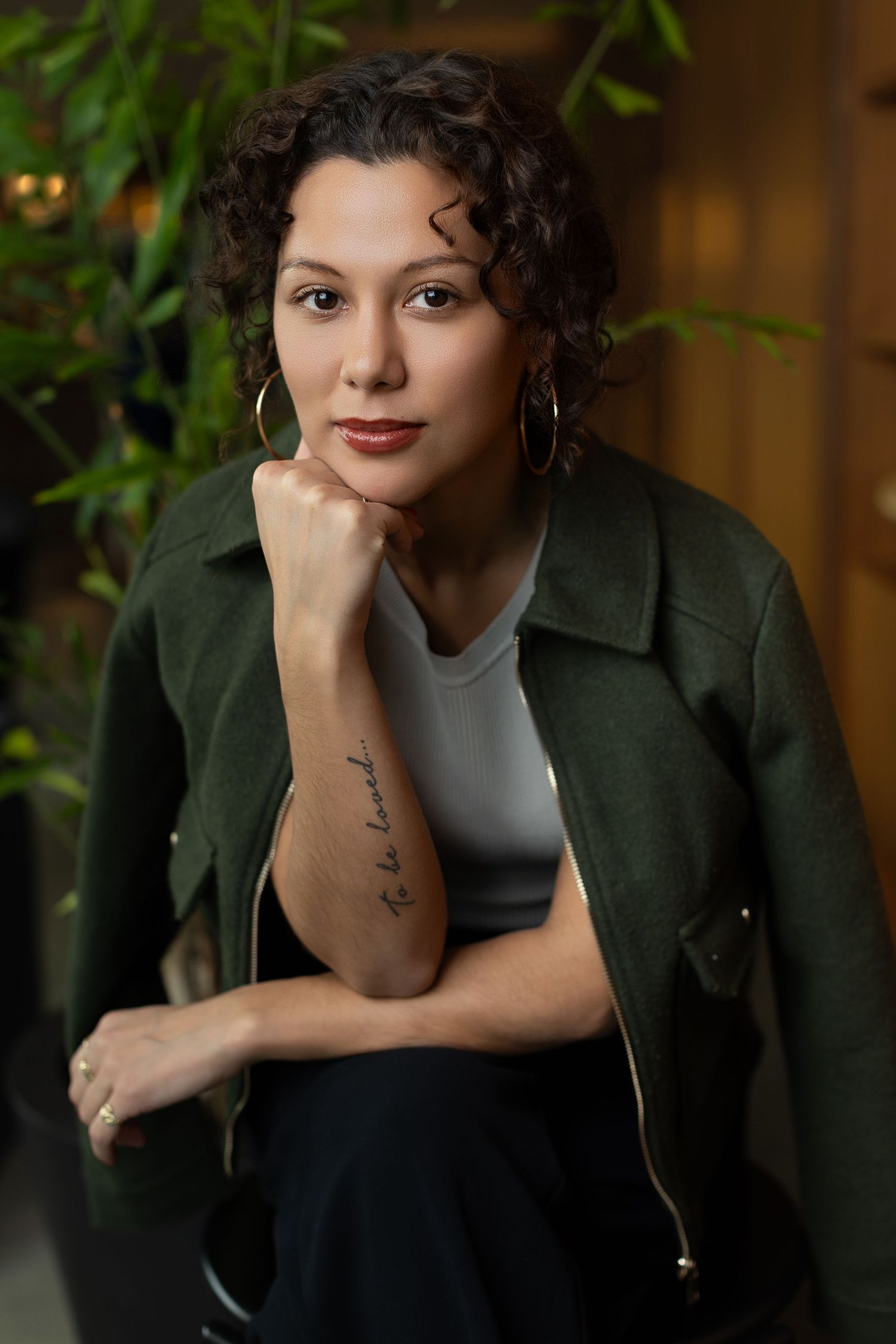 Professional portrait in warm lighting with dark green jacket and white top against foliage background.