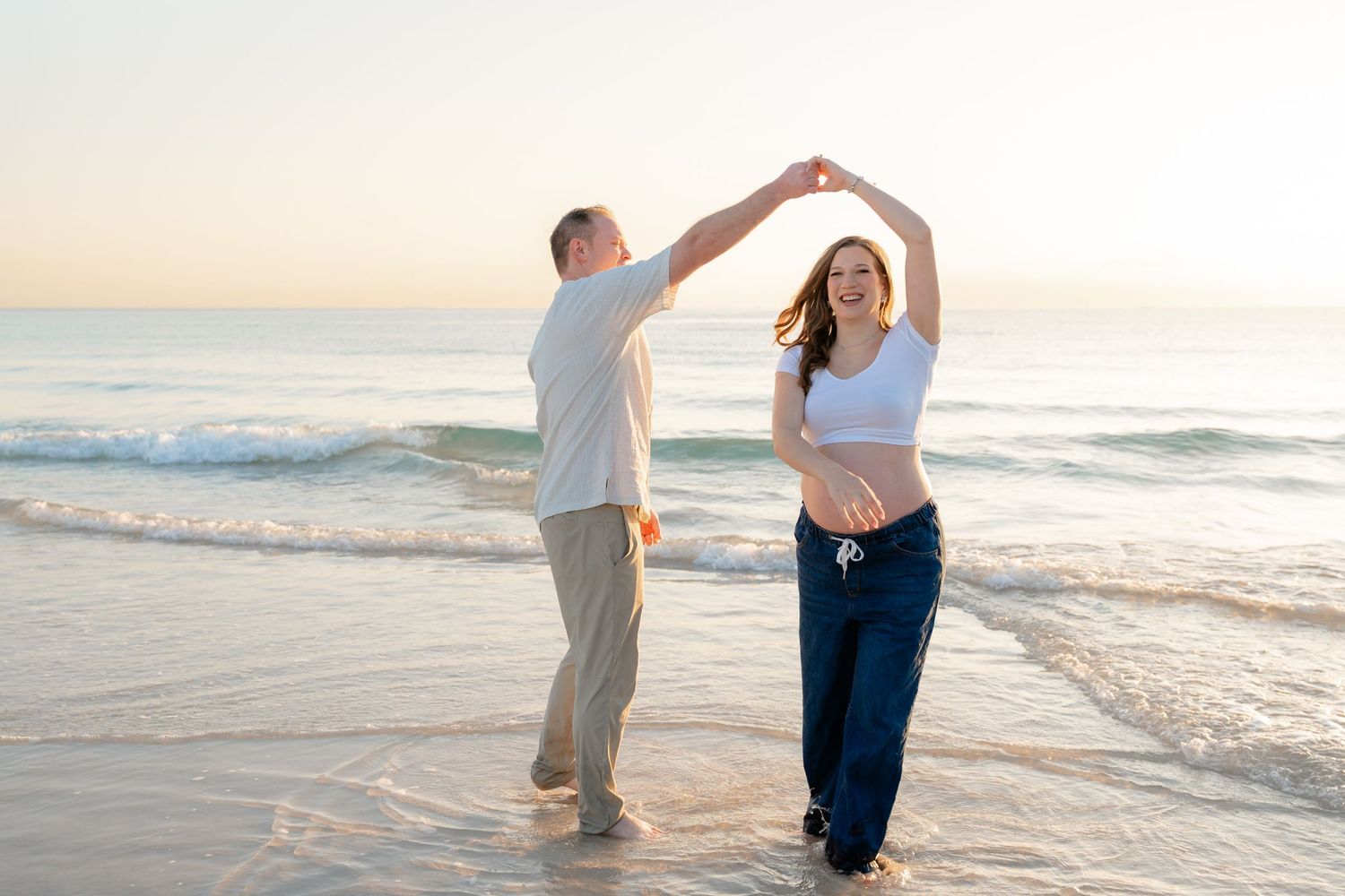 A couple dances together on a sandy beach at sunset with ocean waves in the background.