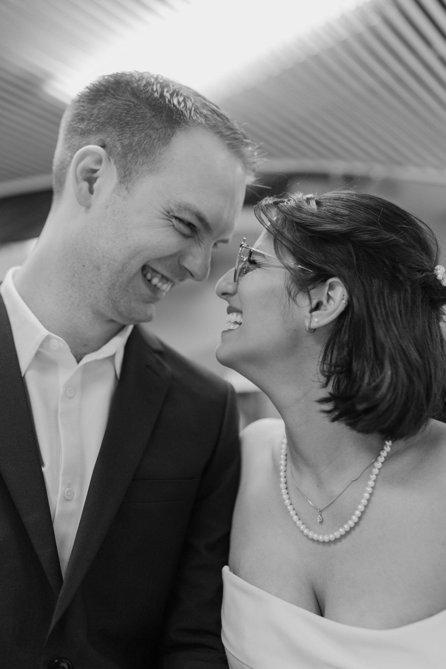 Black and white portrait of laughing newlywed couple touching foreheads, bride wearing pearl necklace.