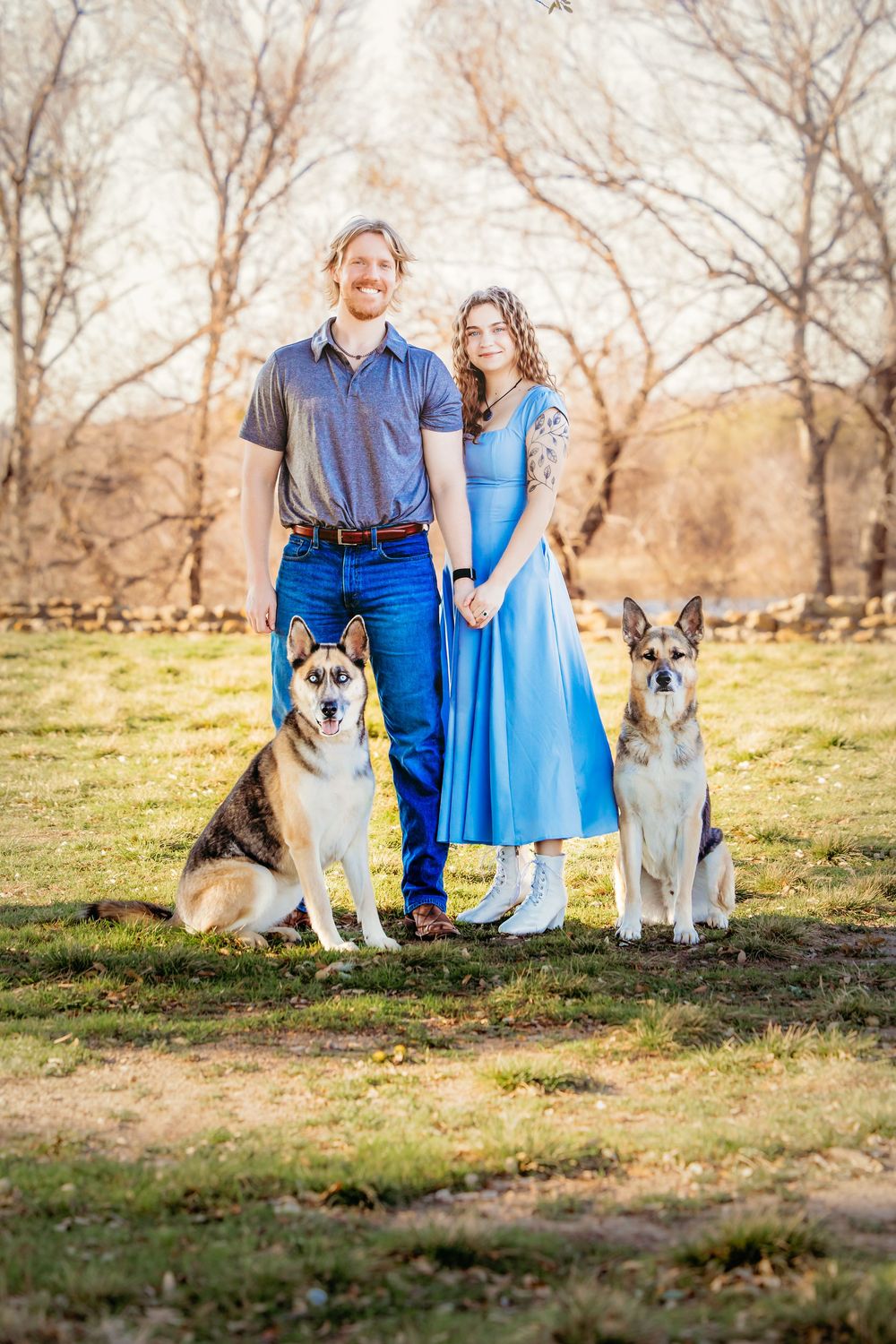 A couple in blue attire poses outdoors with two German Shepherd dogs on a sunny day in a natural setting.