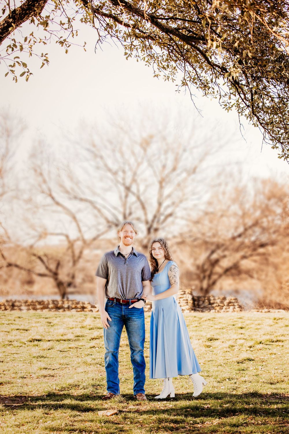 A couple stands together in a sunlit orchard field wearing blue and denim on a warm autumn afternoon.