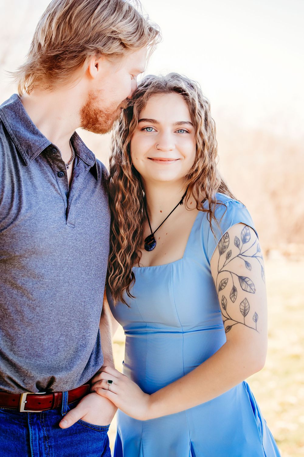 A couple in casual attire share a tender moment outdoors, with her in a blue dress and him in a grey polo shirt.