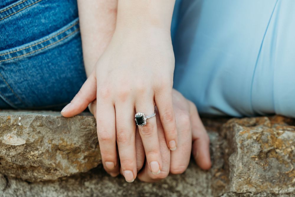 Close-up view of hands with engagement ring resting on rustic stone wall against denim background.