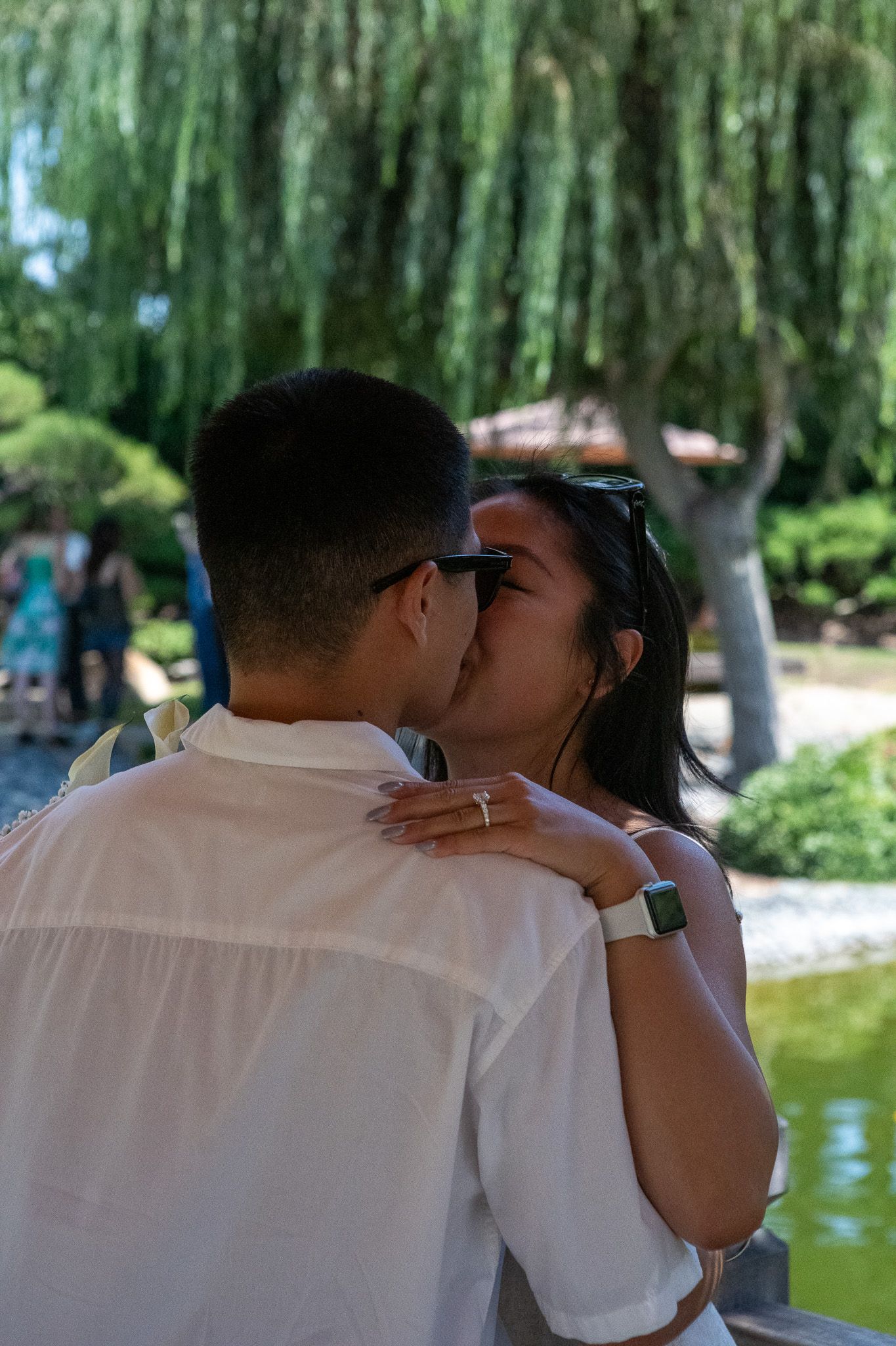 A couple shares a tender moment under willow trees beside a tranquil pond in a serene garden setting.