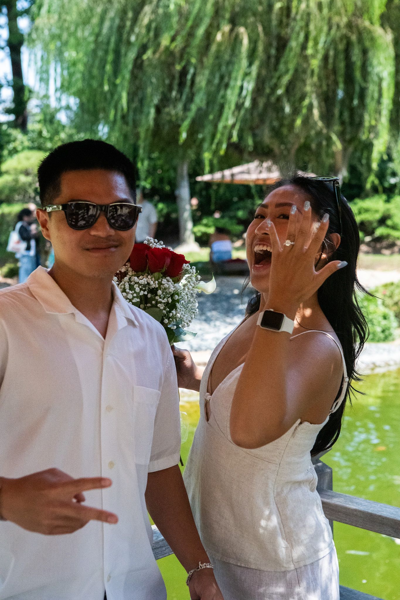 A couple laughs together while enjoying a sunny day by a pond with willow trees in the background.