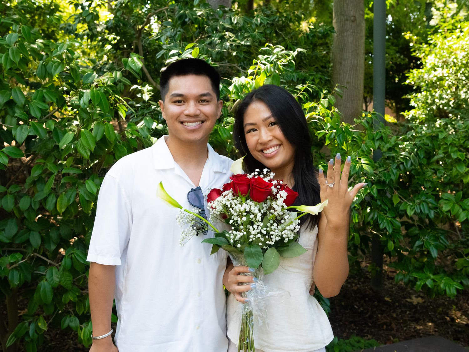A couple poses outdoors in white outfits with a bouquet of red flowers against a leafy green background.