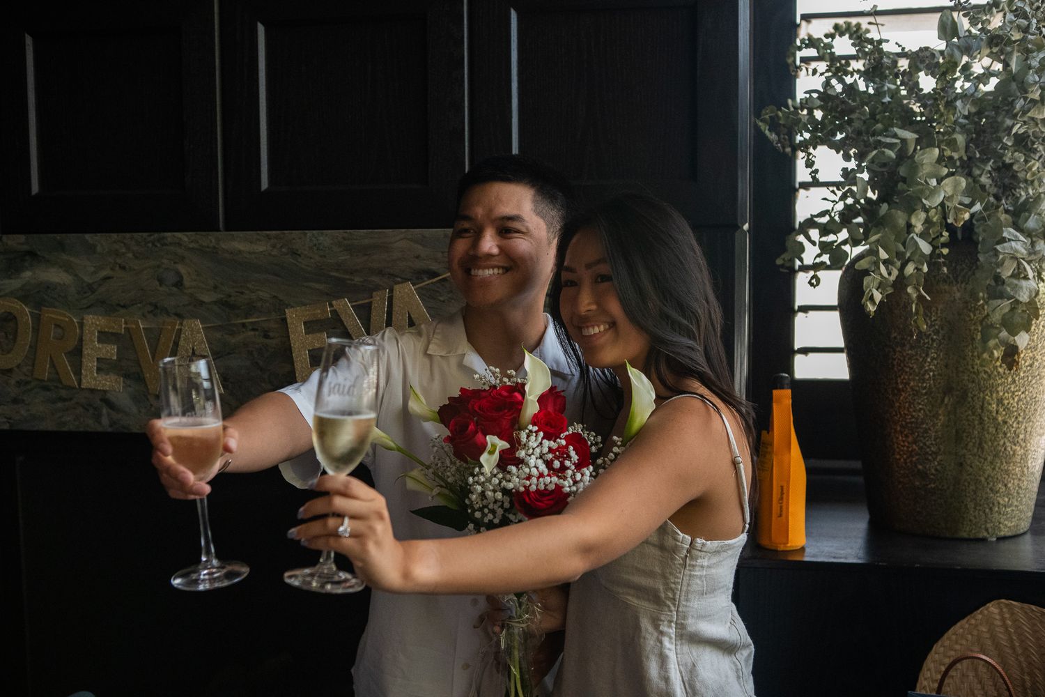 A couple celebrates with champagne glasses and a bouquet of red roses at a dimly lit restaurant.