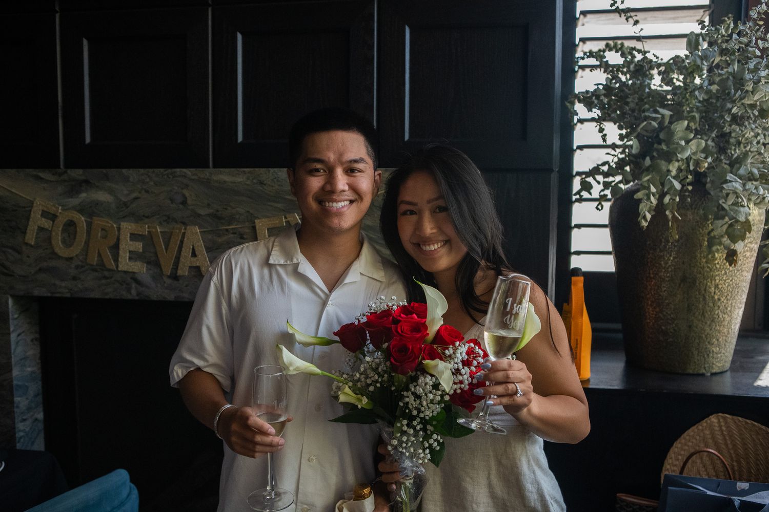 A couple smiles while holding drinks and a red floral bouquet in front of a rustic wooden sign.