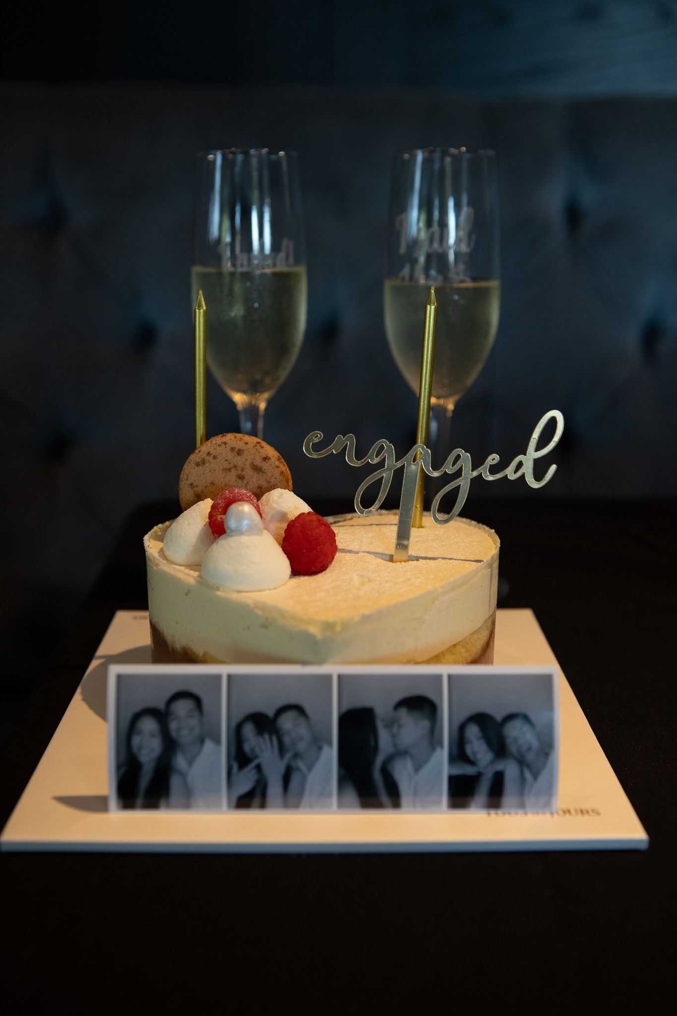 An engagement celebration dessert with champagne glasses and photo strip displayed on a white cake plate.