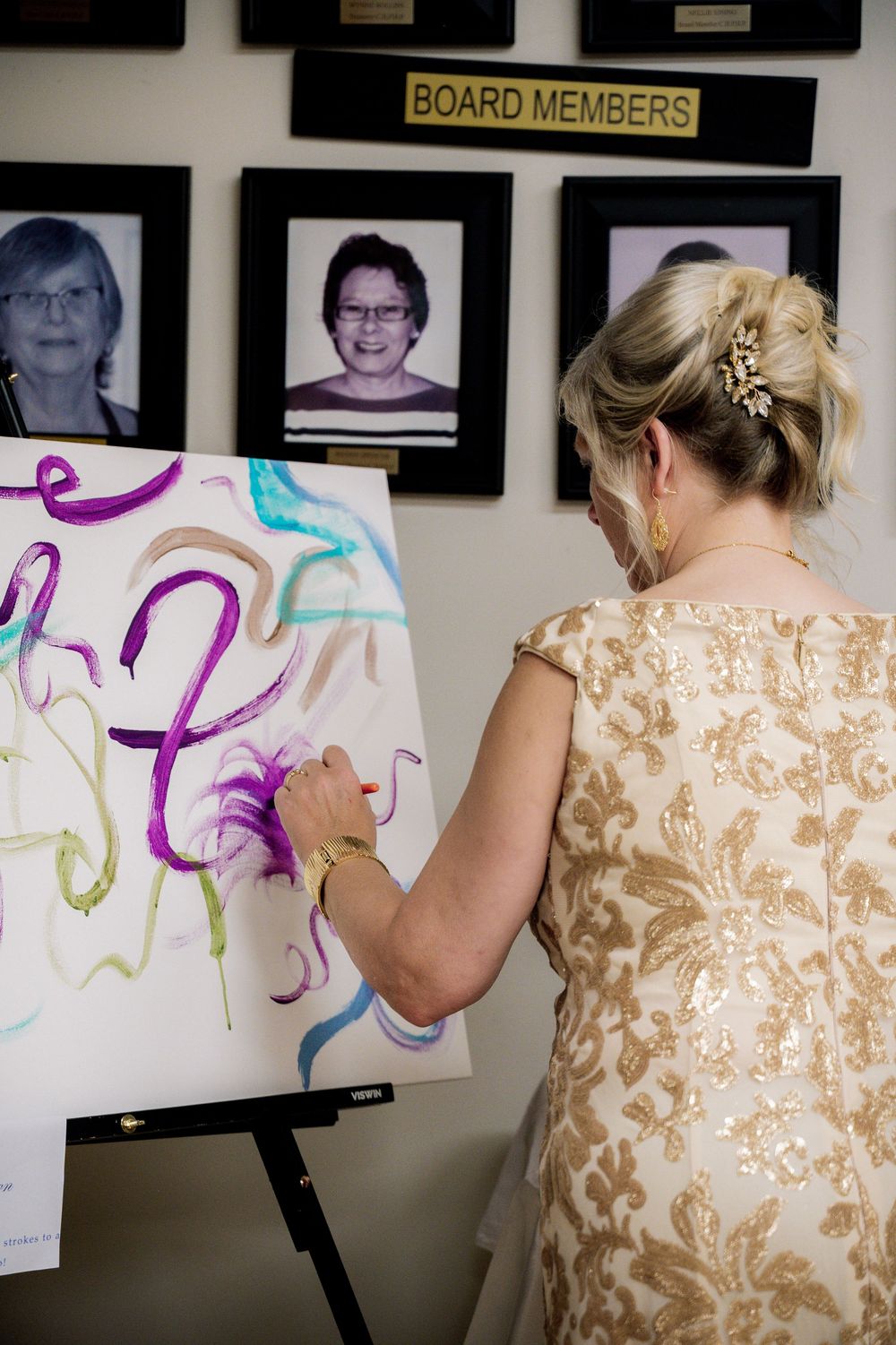 A person in a floral dress draws colorful artwork on white paper displayed on a wall with board member photos.