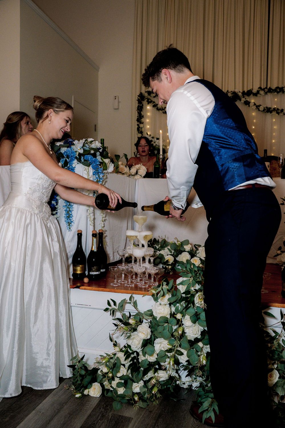 Wedding couple pours champagne in a tower of glasses surrounded by elegant white floral decorations at their reception.