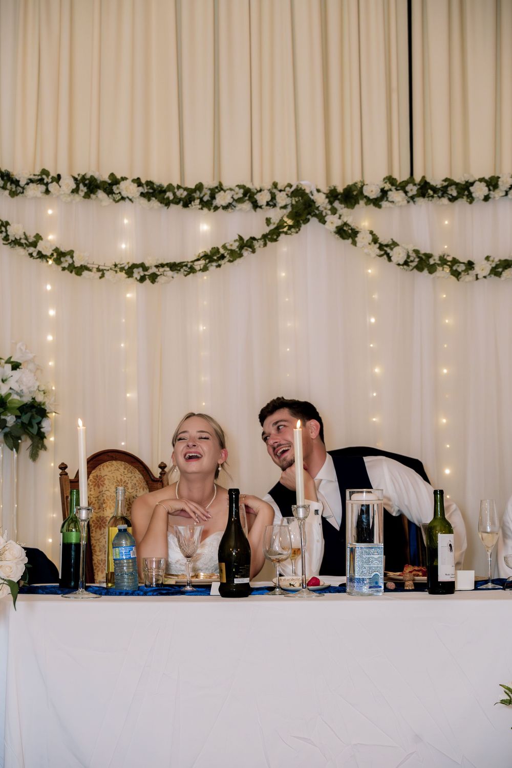 Wedding reception guests seated at a long table with white drapes and twinkling lights in the background.