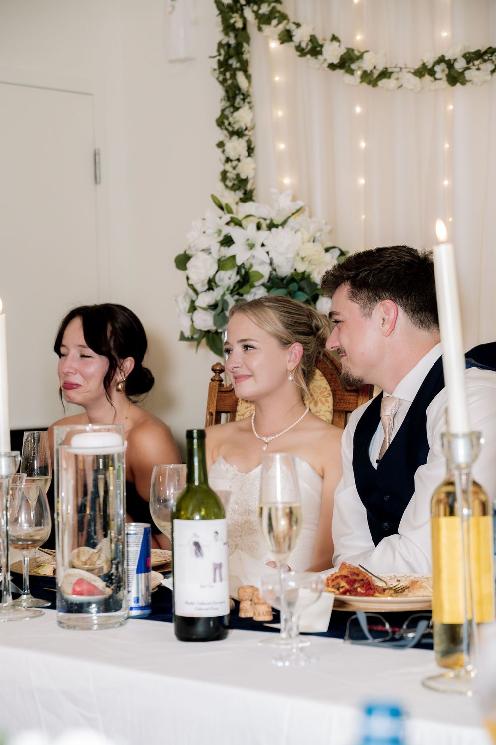 Wedding party guests seated at a formal dining table decorated with greenery and candles enjoy conversation and drinks.