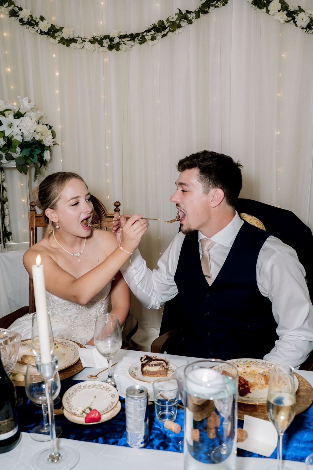 A couple playfully feeds each other cake at their wedding reception table decorated with greenery and twinkle lights.