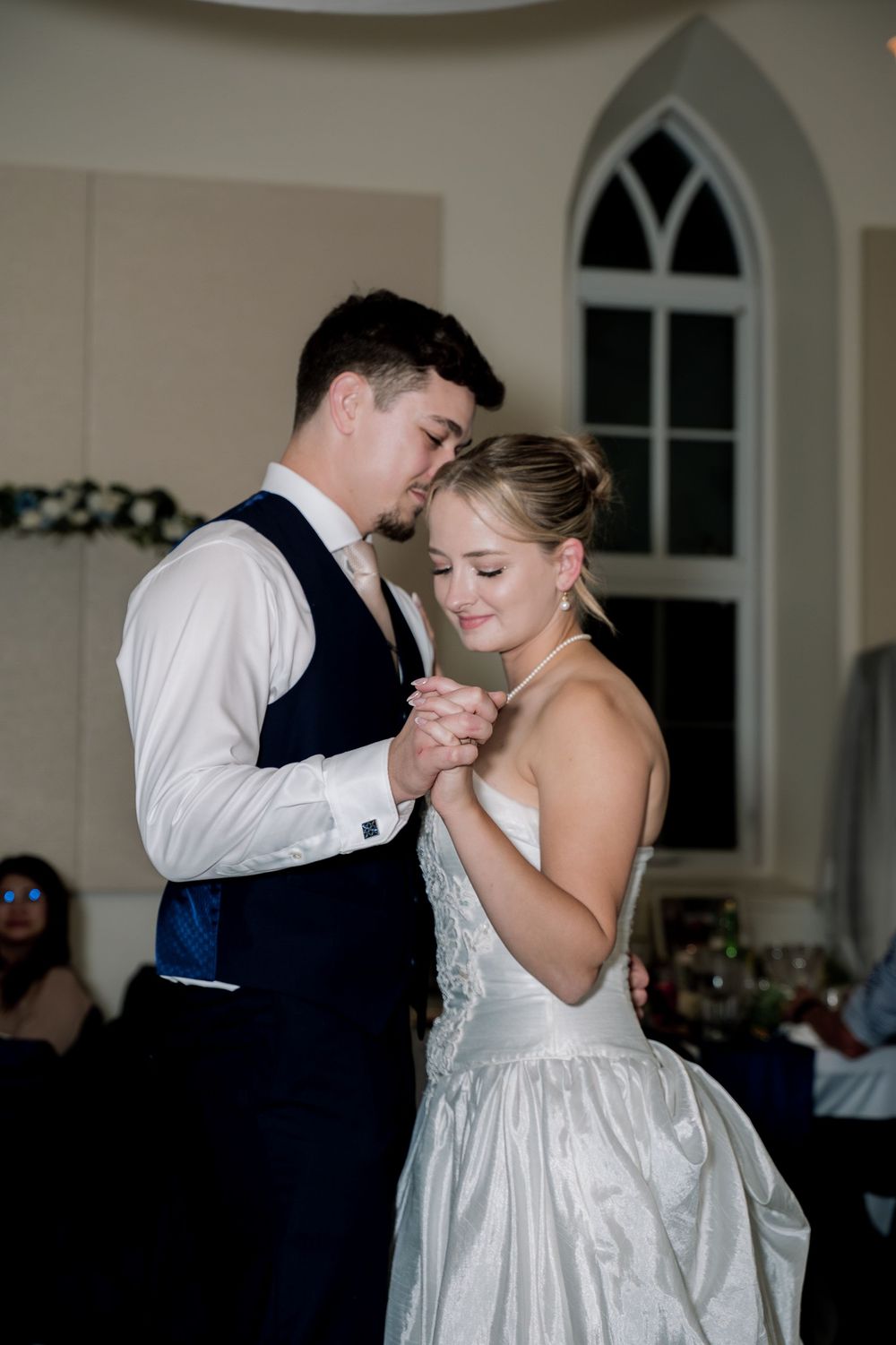 A couple shares an intimate dance moment at a wedding reception in an elegant white venue with arched windows.