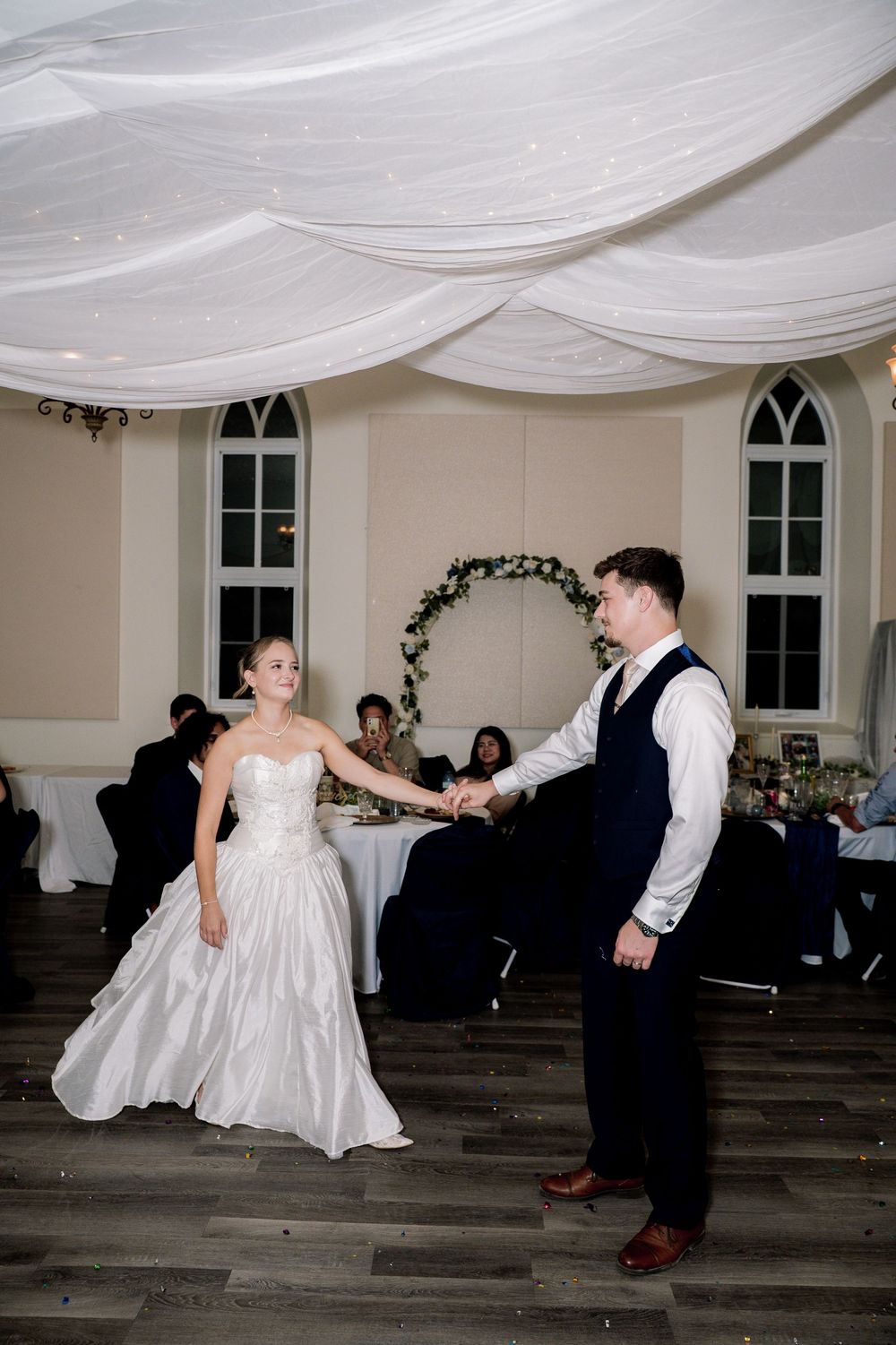 A couple dances together at their wedding reception under elegant white fabric draping and arched windows.