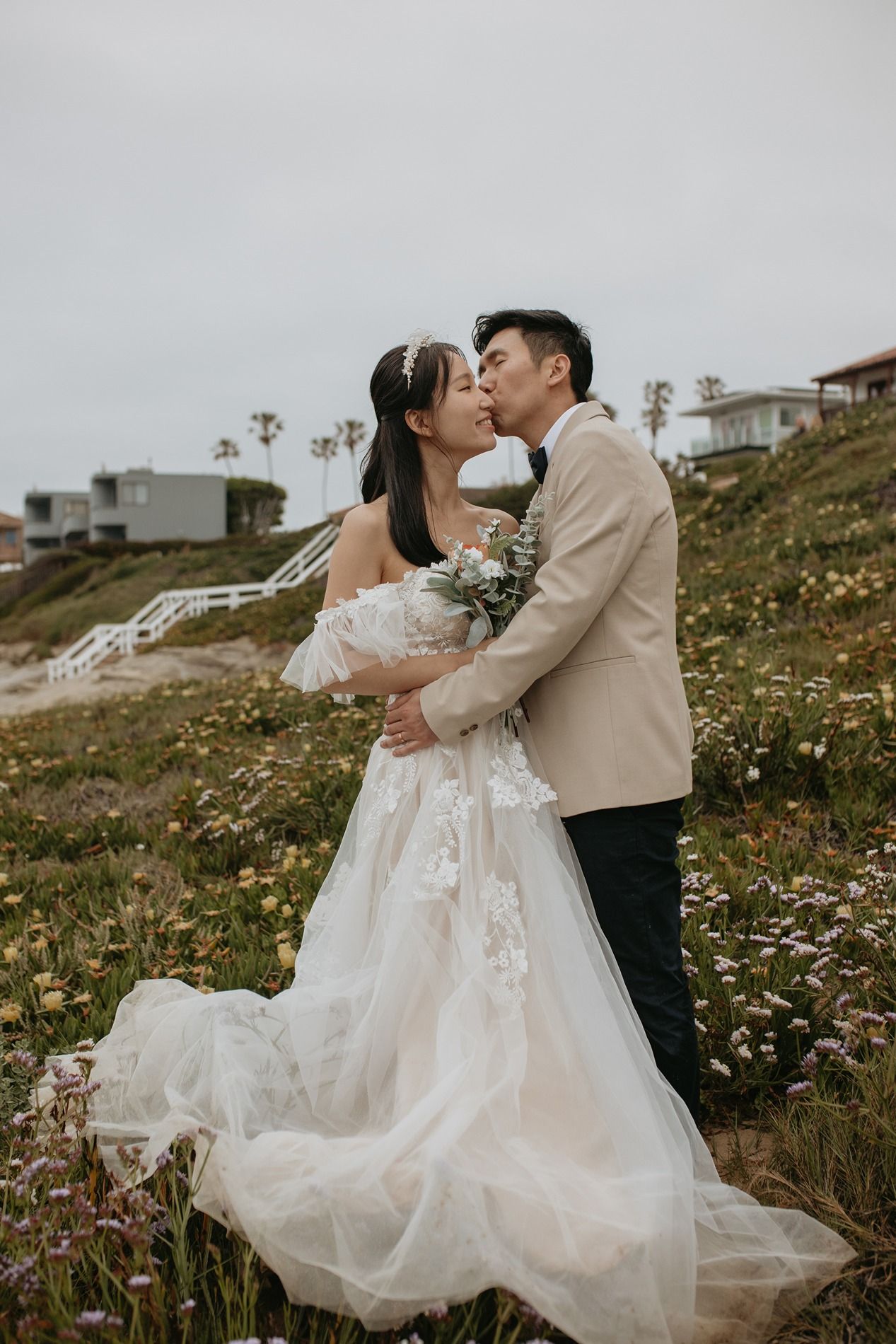 A couple shares a romantic kiss in a field of wildflowers while the bride wears a flowing lace wedding gown at Windansea Beach in La Jolla.