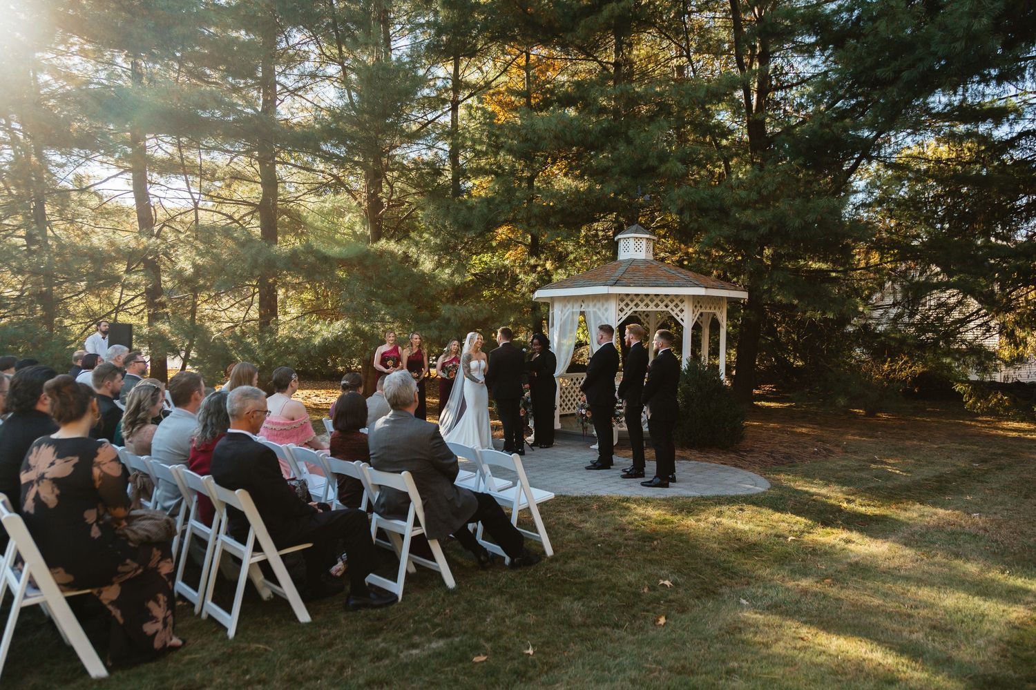 Outdoor wedding ceremony at a gazebo surrounded by tall trees with guests seated on white chairs.