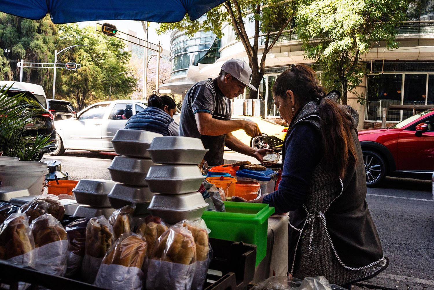 A street vendor prepares and serves food from a mobile cart on a sunny urban sidewalk.