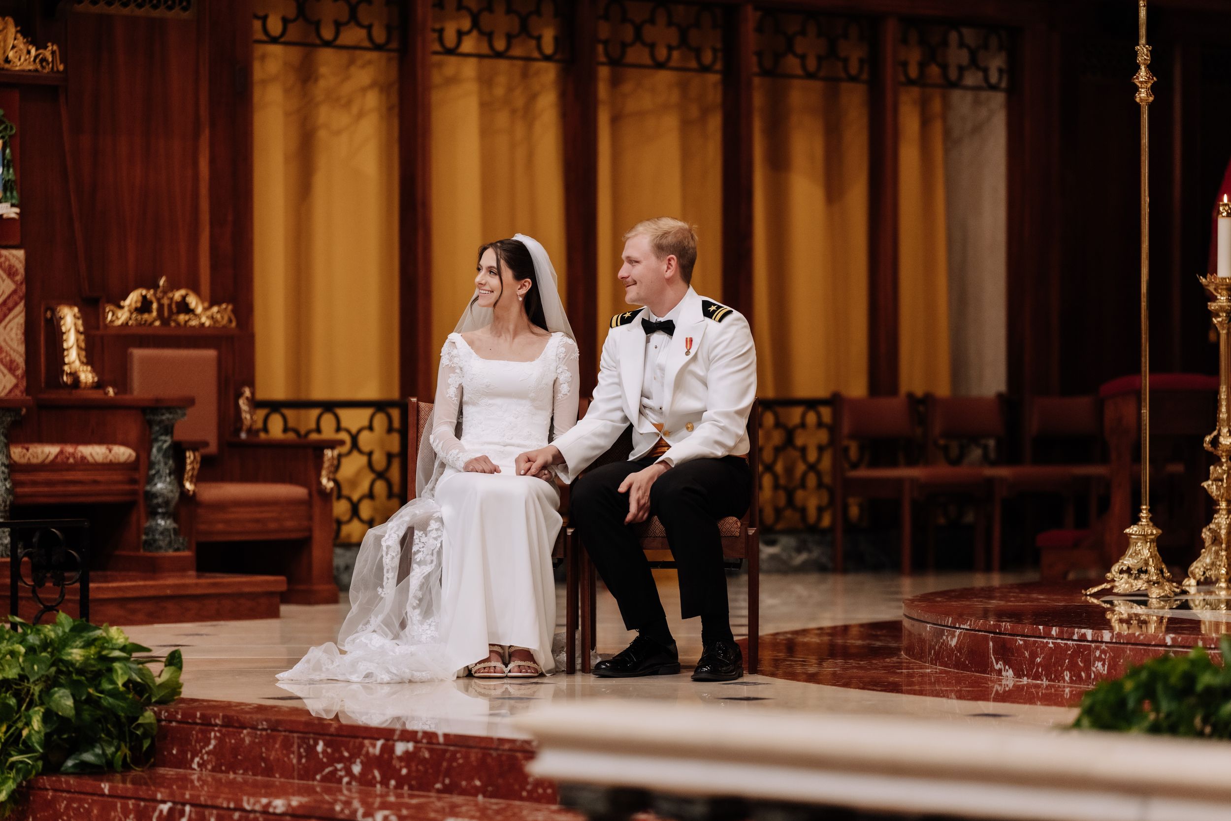 Wedding couple seated on red carpeted steps inside a cathedral with warm lighting and wooden details.