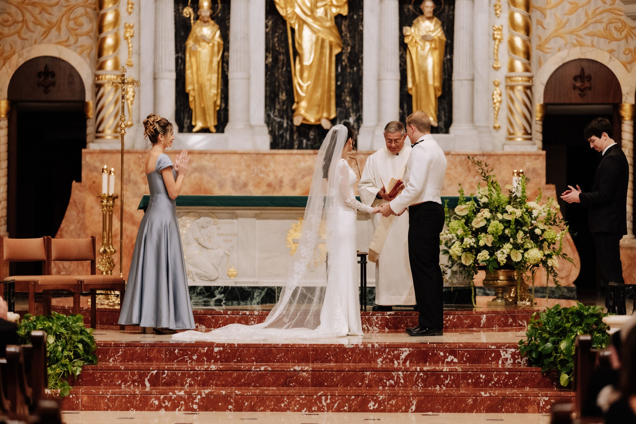 Traditional wedding ceremony taking place at an ornate church altar with golden statues and floral arrangements.