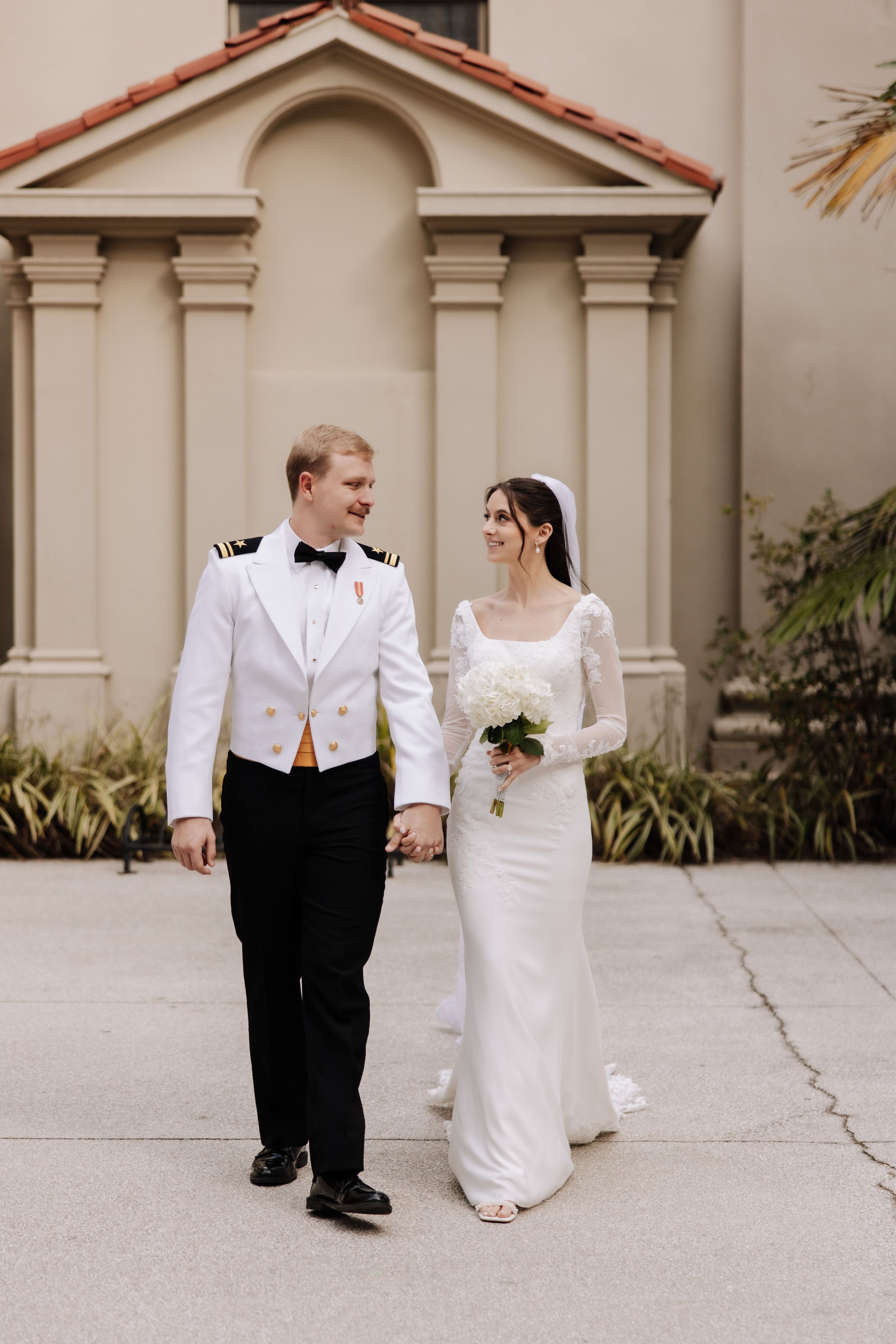 Newlywed couple walking and holding hands along a concrete path beside a Mediterranean-style building.