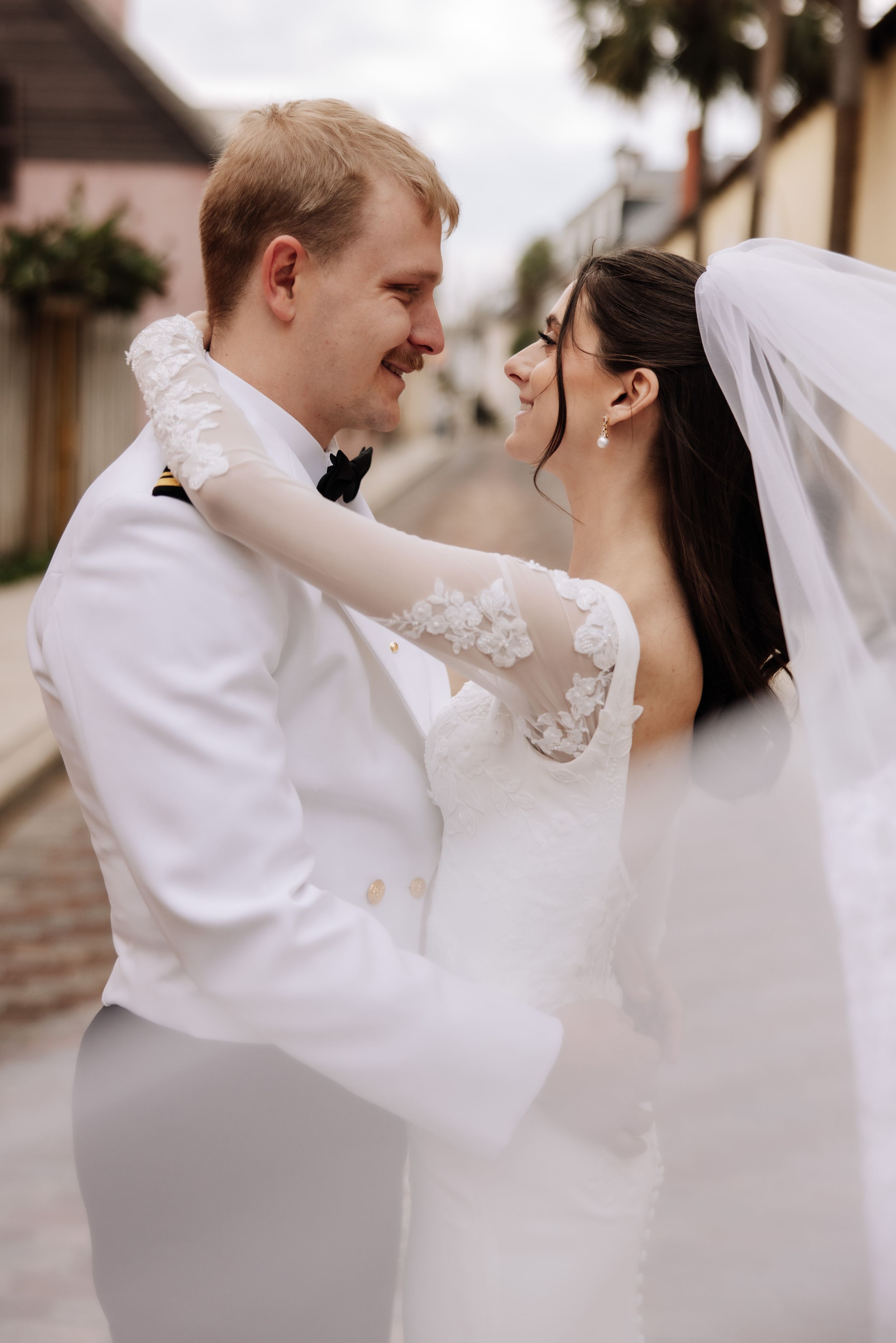 Happy couple in wedding attire dancing and twirling together outside a beige building.