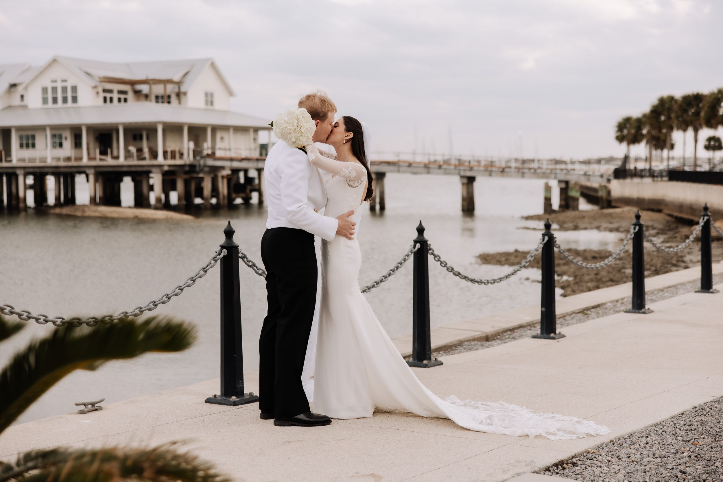 A romantic wedding couple embraces on a waterfront boardwalk with a coastal house and harbor in the background.