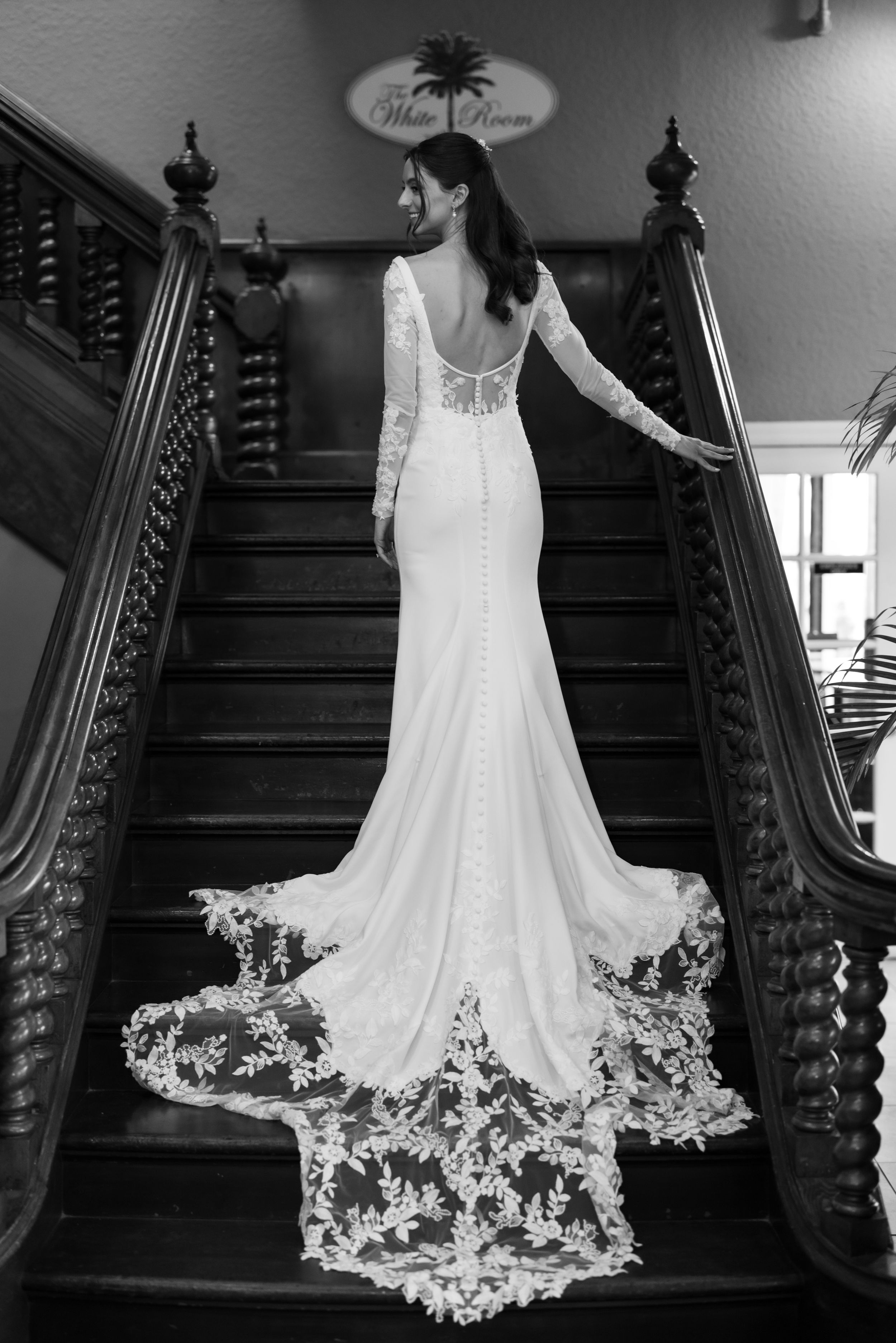 A stunning white wedding dress with intricate lace train displayed on a dark wooden staircase in black and white.