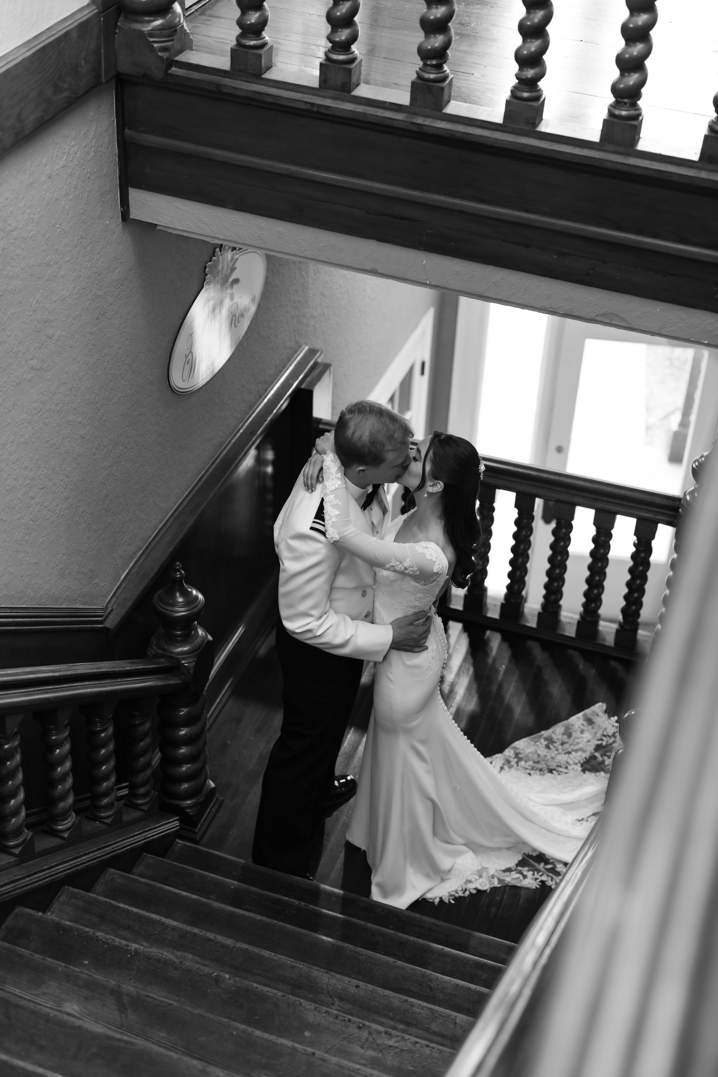 A romantic black and white photograph of a couple embracing on an elegant wooden staircase with ornate balusters.