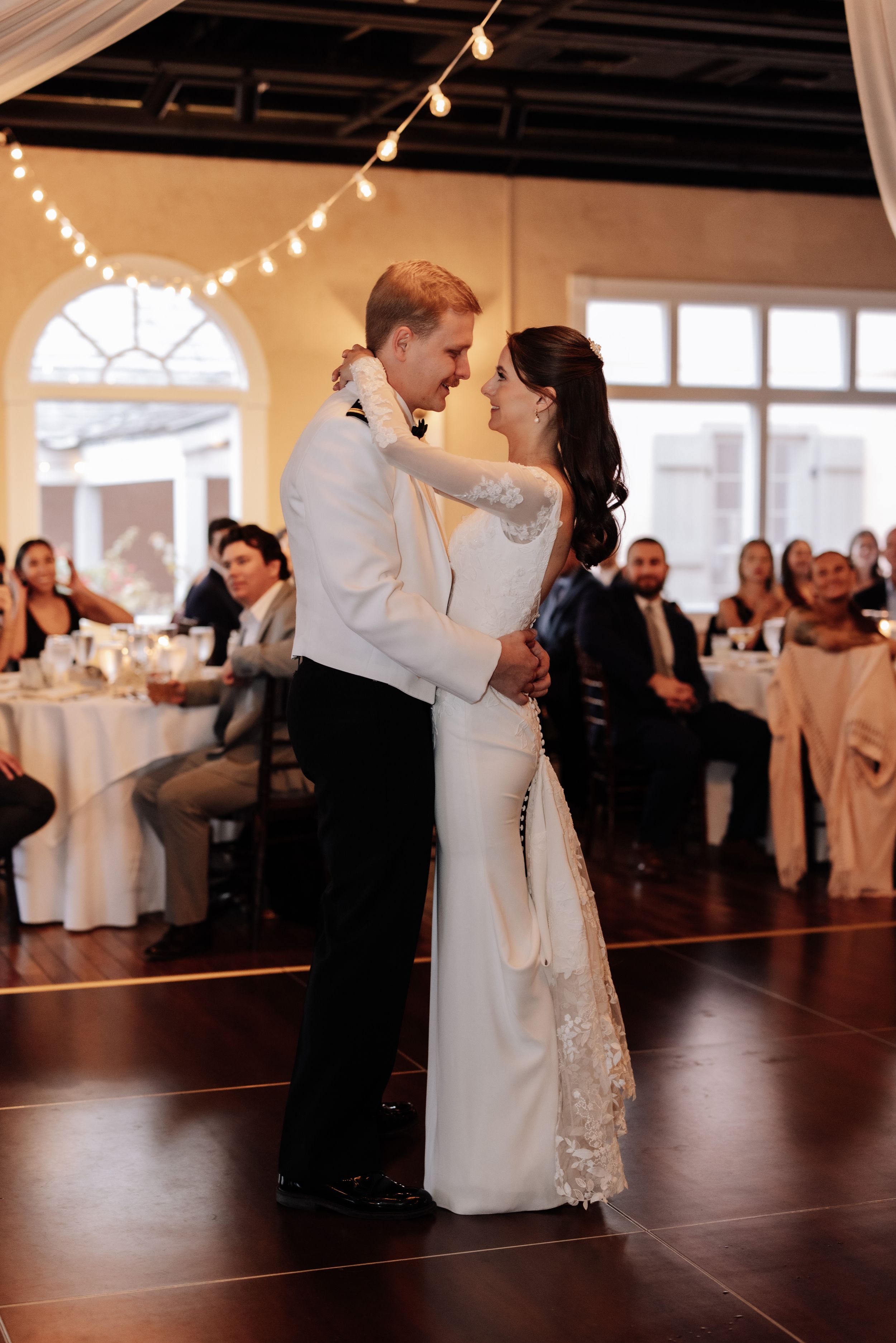A couple shares their first dance at their wedding reception under string lights in an elegant venue with dark floors.