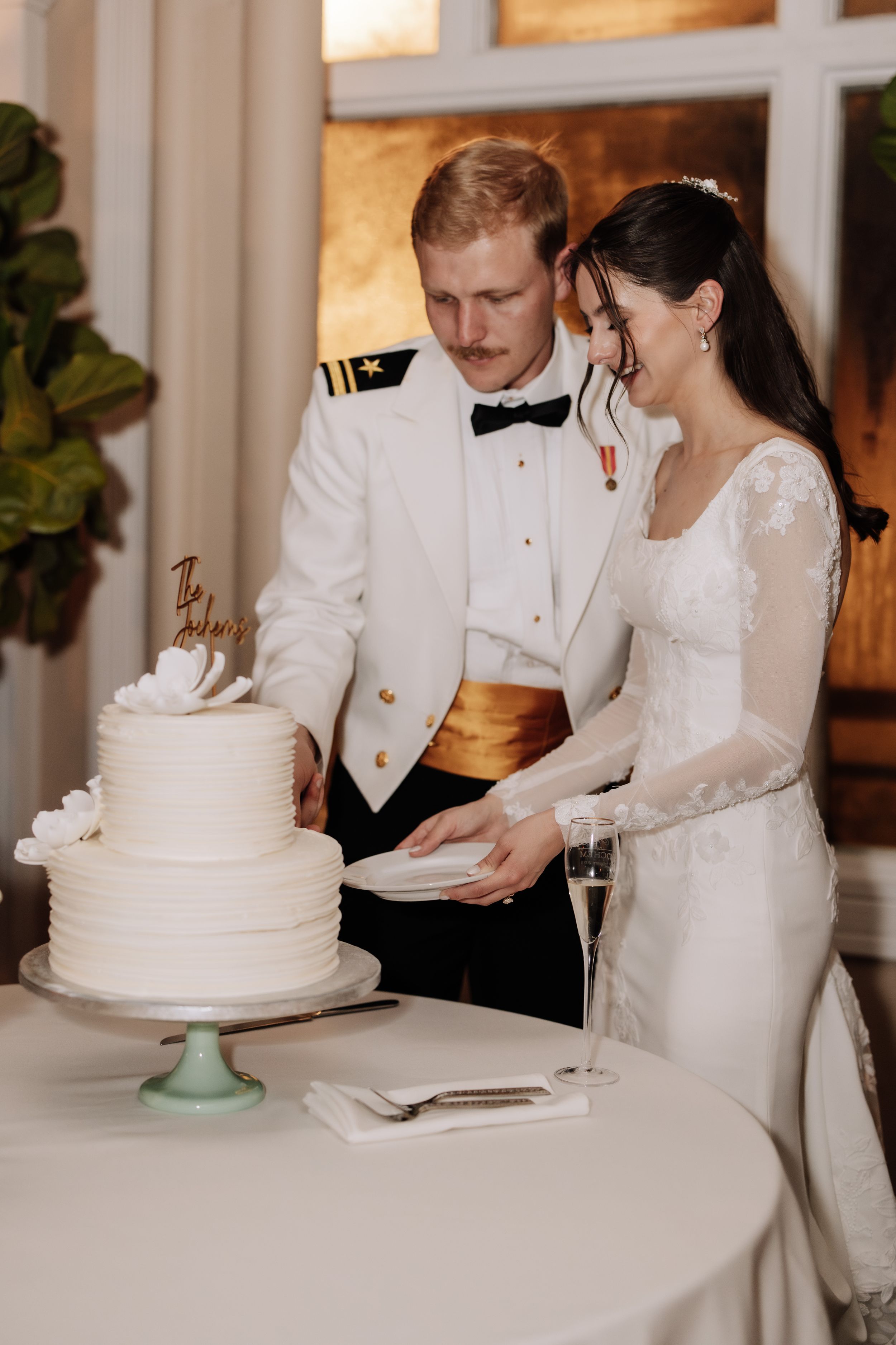 A bride and groom in formal attire sign their marriage license at a white-clothed table next to their wedding cake.