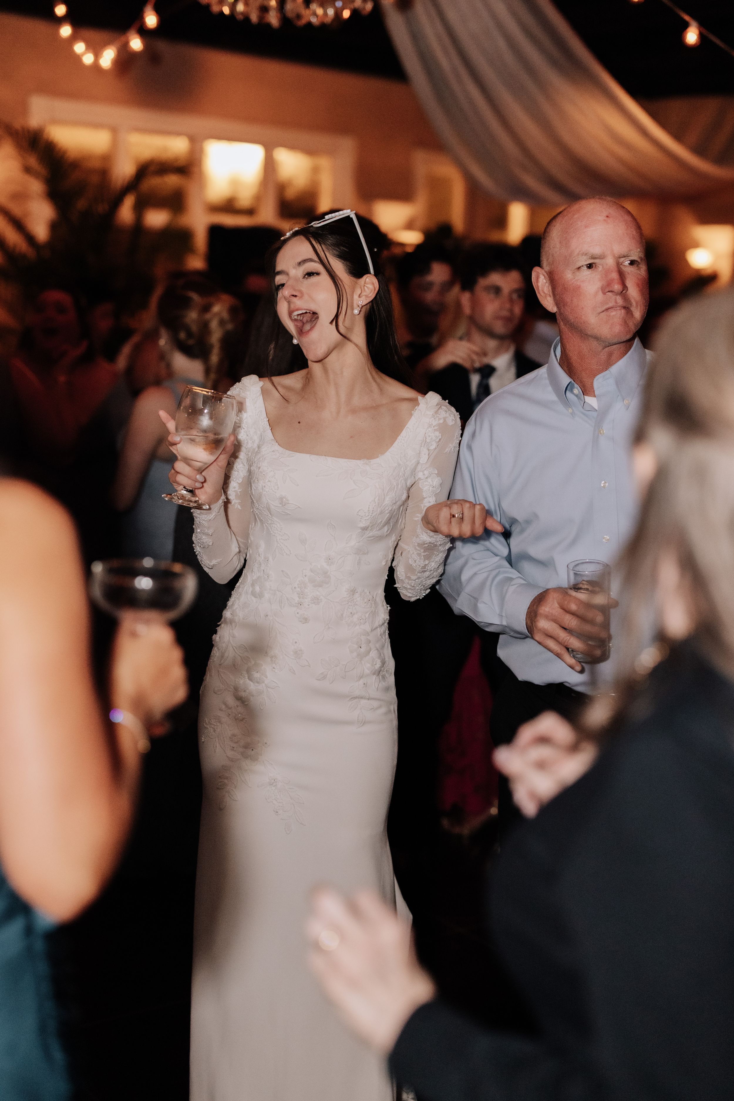 People dancing and celebrating at a dimly lit wedding reception with string lights overhead and festive atmosphere.