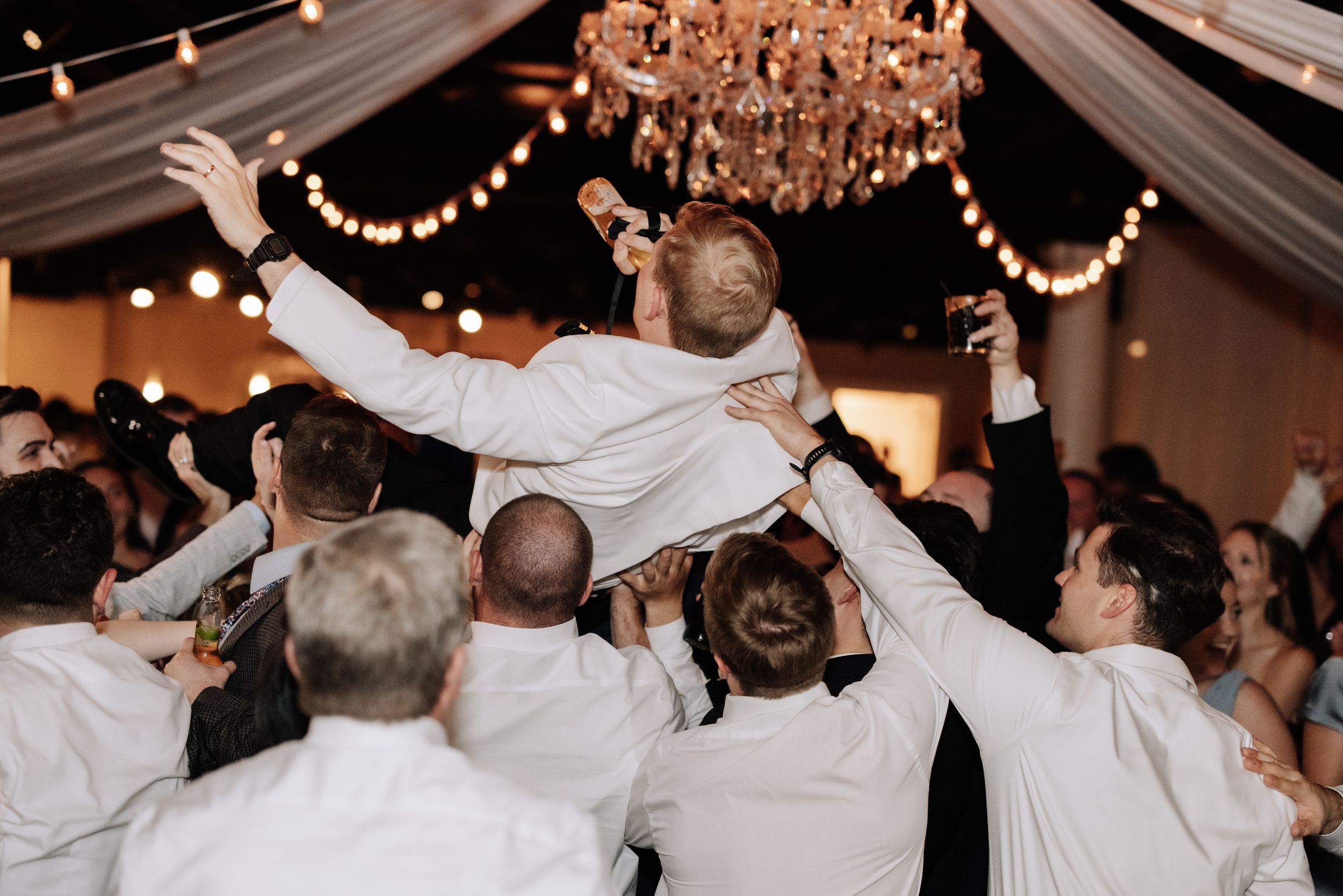 Wedding guests celebrate on the dance floor with energetic dance moves under sparkling chandeliers and string lights.