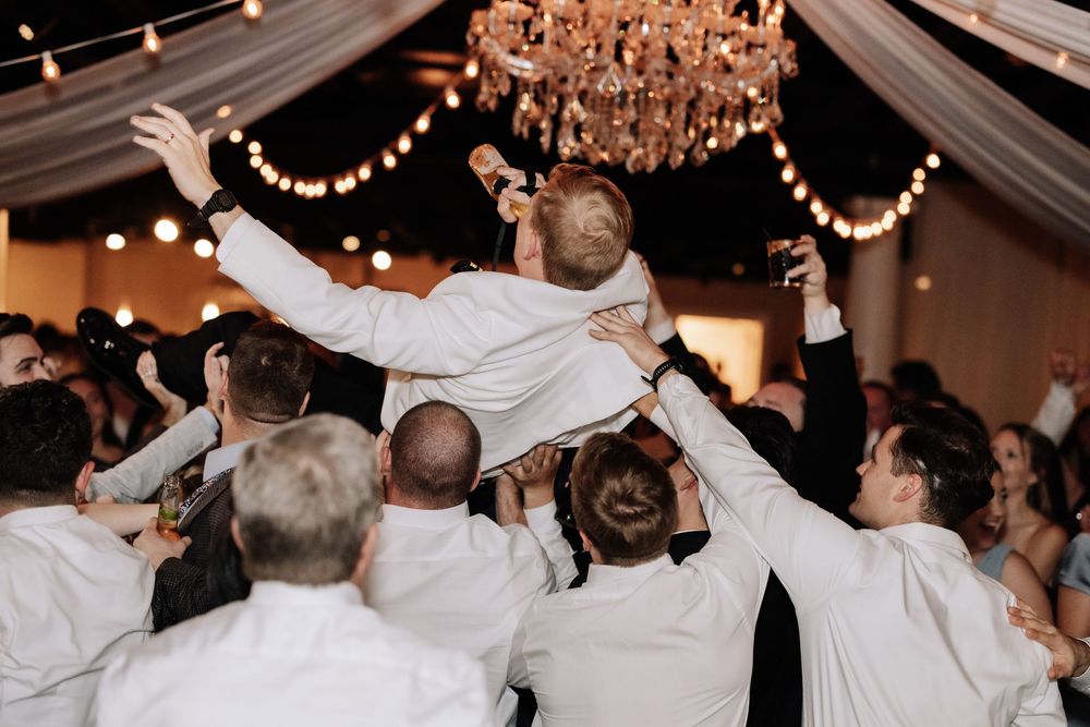 Wedding guests celebrate on the dance floor with energetic dance moves under sparkling chandeliers and string lights.