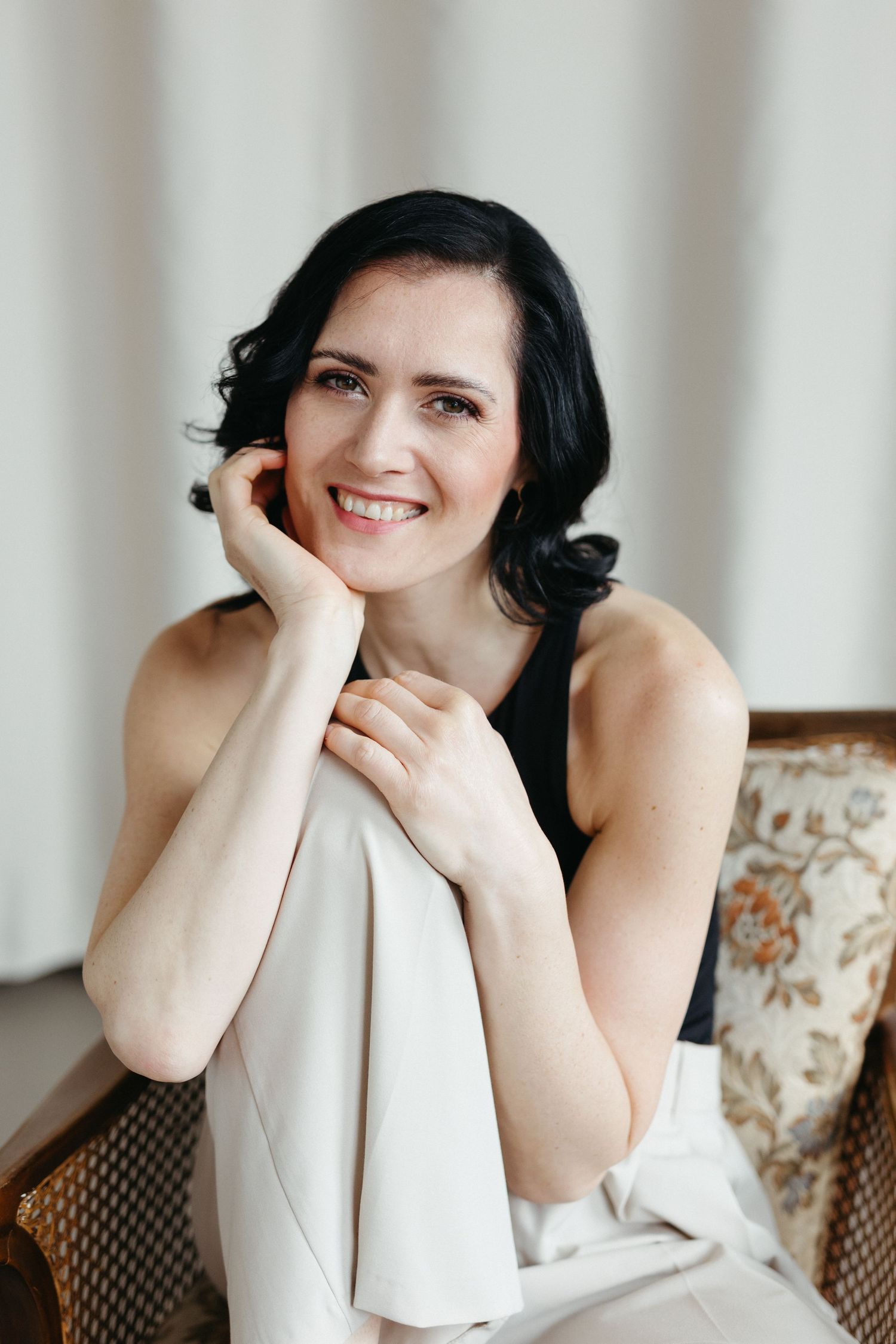 A person with dark hair smiles while resting on a chair in a bright indoor setting.