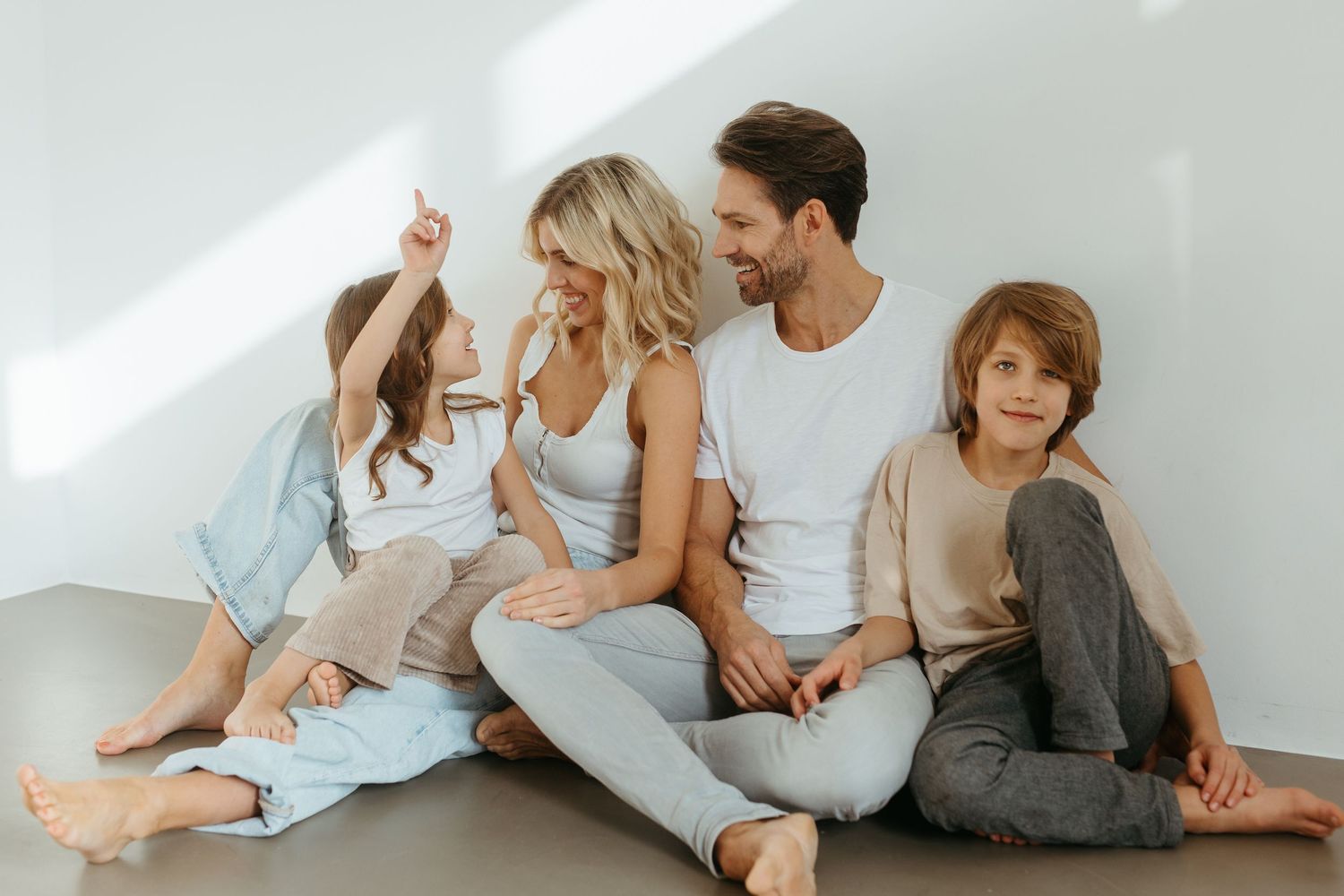 A family in casual clothing sits together on the floor against a white wall in natural lighting.