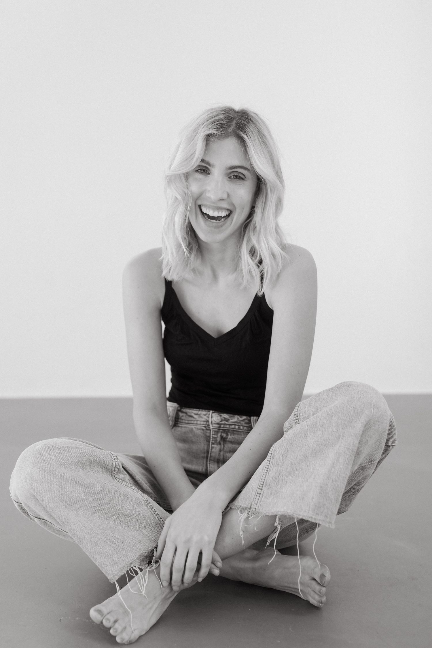 Black and white portrait of someone sitting cross-legged in casual attire with a joyful expression and blonde wavy hair.