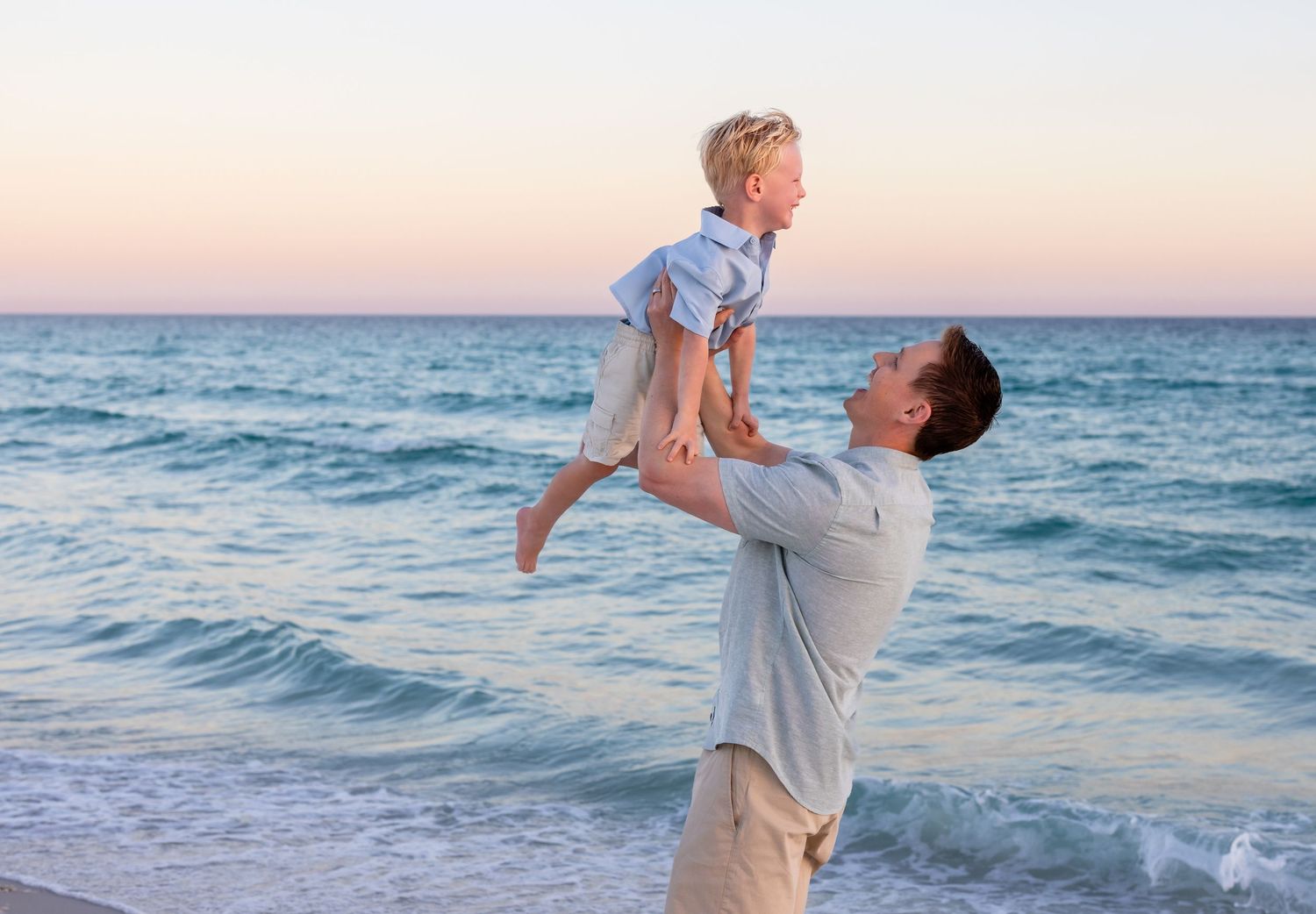 A parent lifts a child playfully in the air while standing on a beach at sunset with ocean waves in the background.