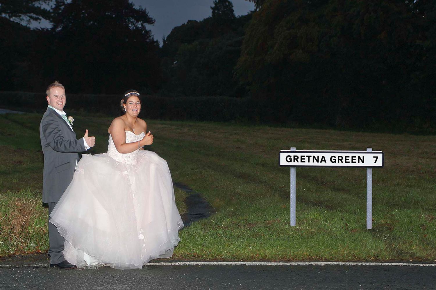A bride and groom stand together on a roadside near a sign for Green Green as they hitch to Gretna for an elopement wedding