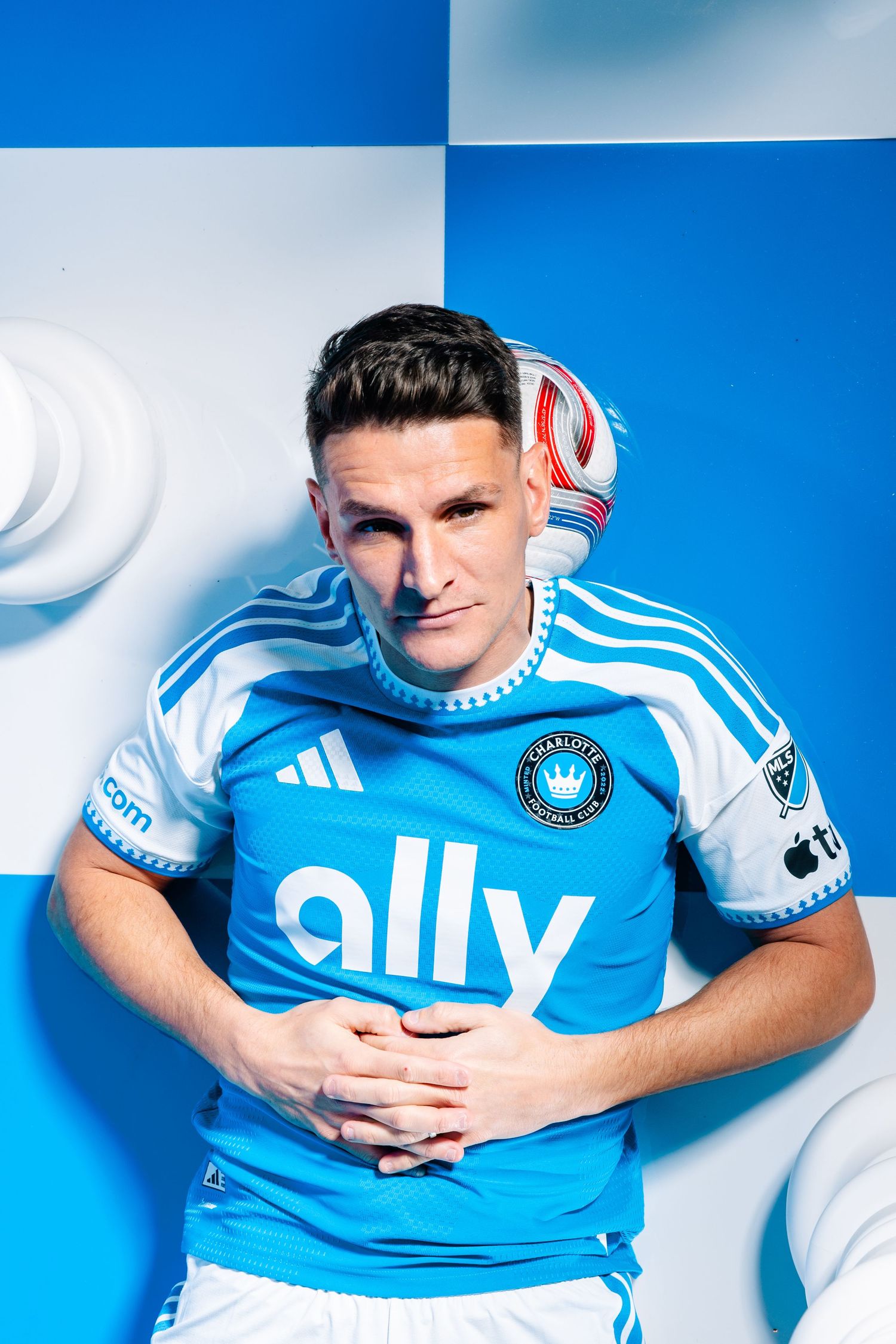 Charlotte FC player poses in blue and white Adidas ally jersey with soccer ball balanced on head.
