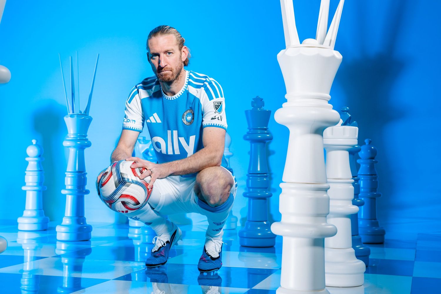 Charlotte FC player crouching with soccer ball among giant chess pieces in blue studio.