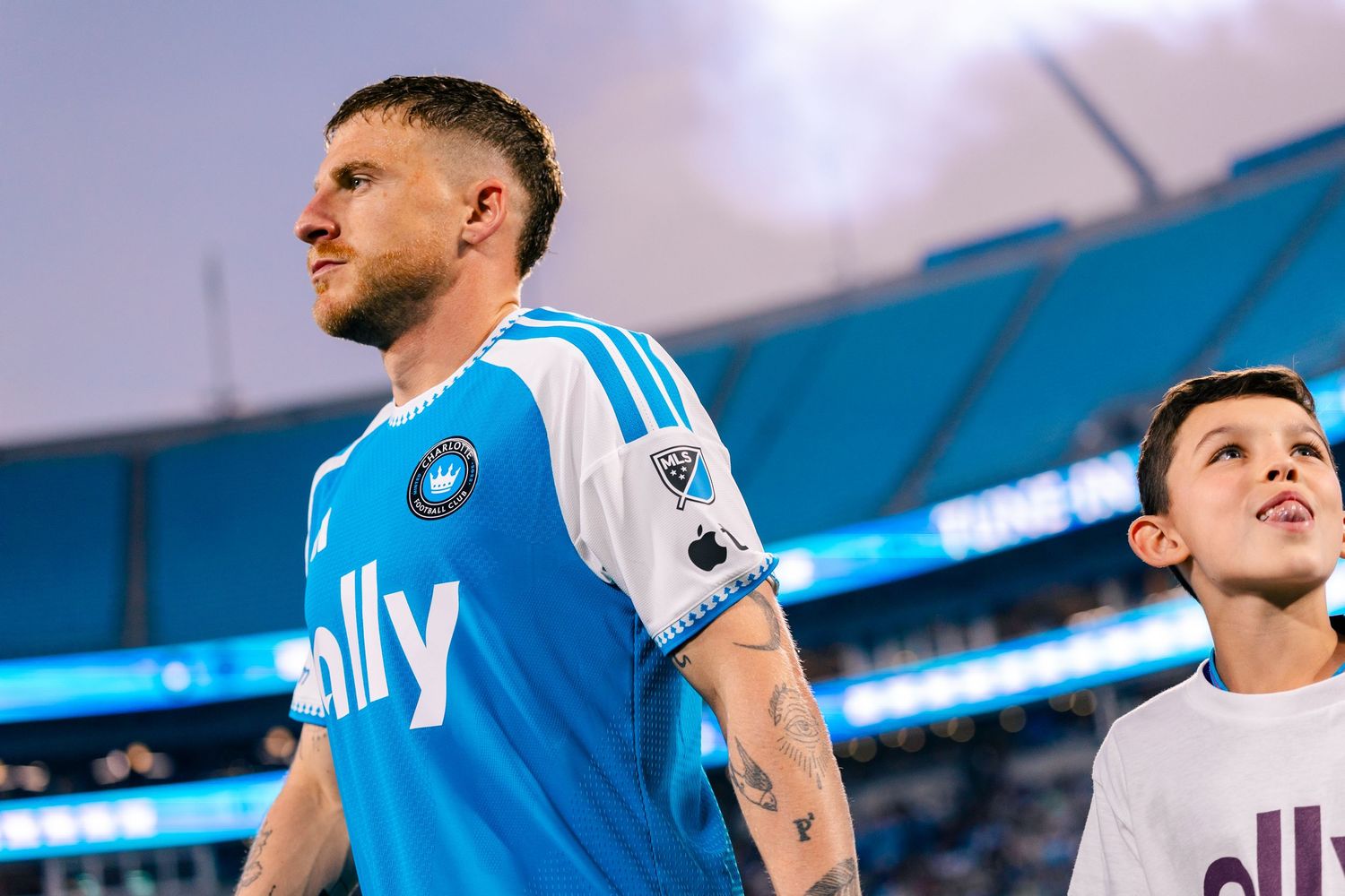 Charlotte FC player in blue ally jersey walks onto field with young mascot during match.