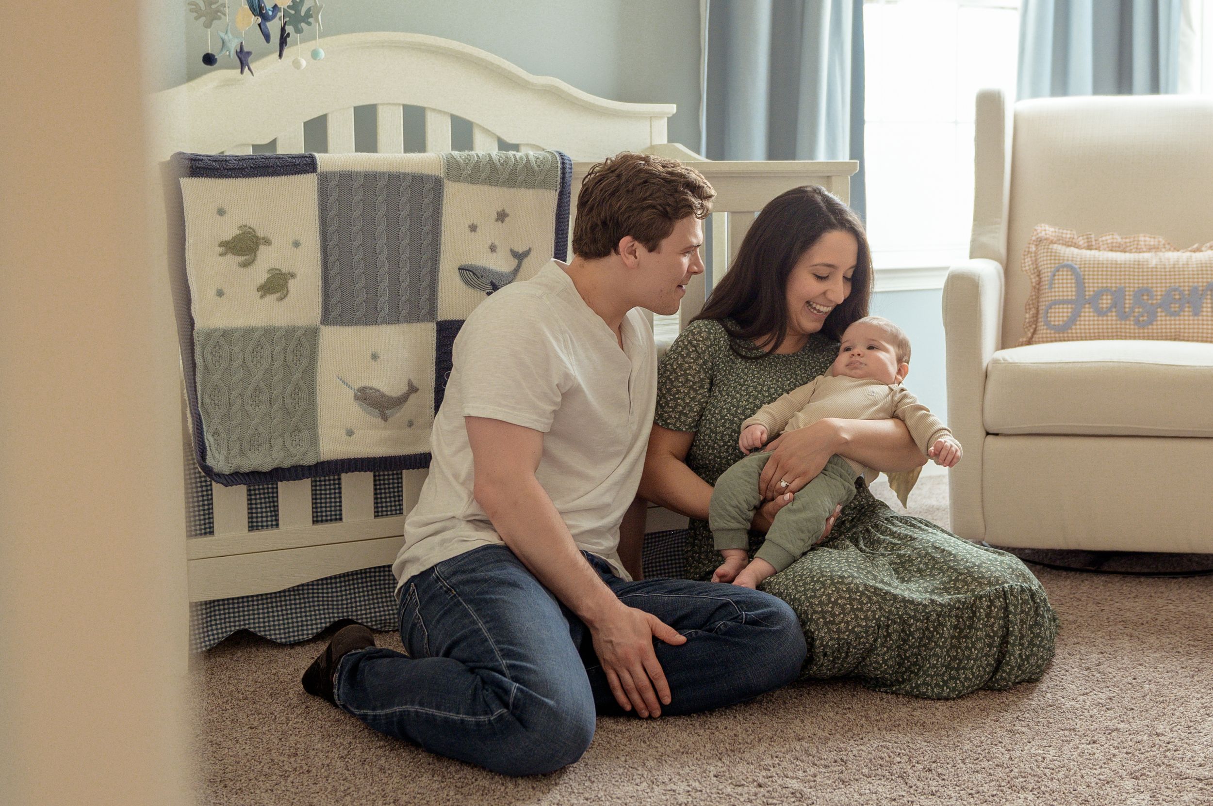 A young couple sitting on the floor with a newborn baby in a cozy nursery setting.