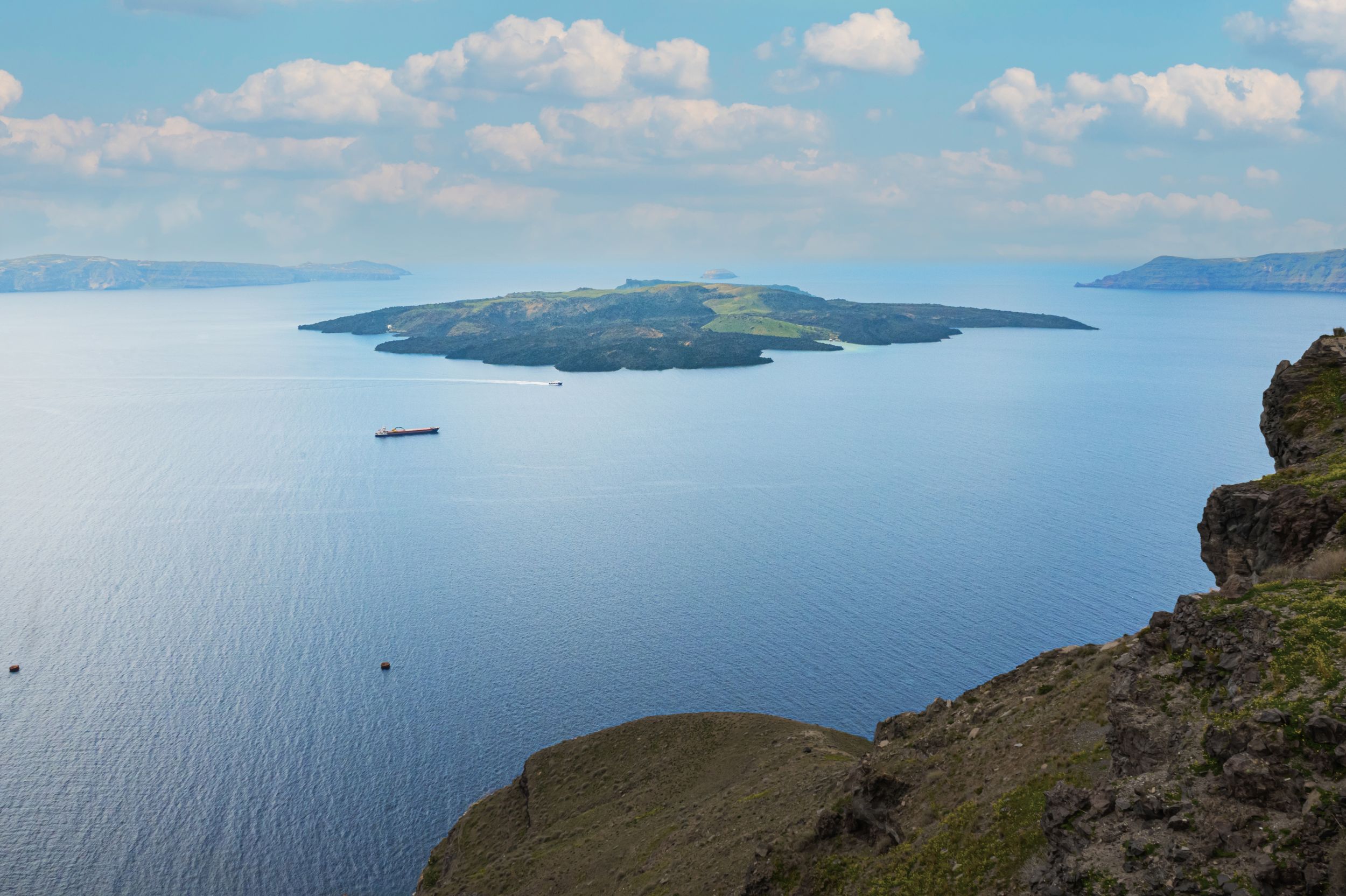 Scenic coastal landscape with dramatic cliffs overlooking a calm blue ocean and distant islands under puffy white clouds.