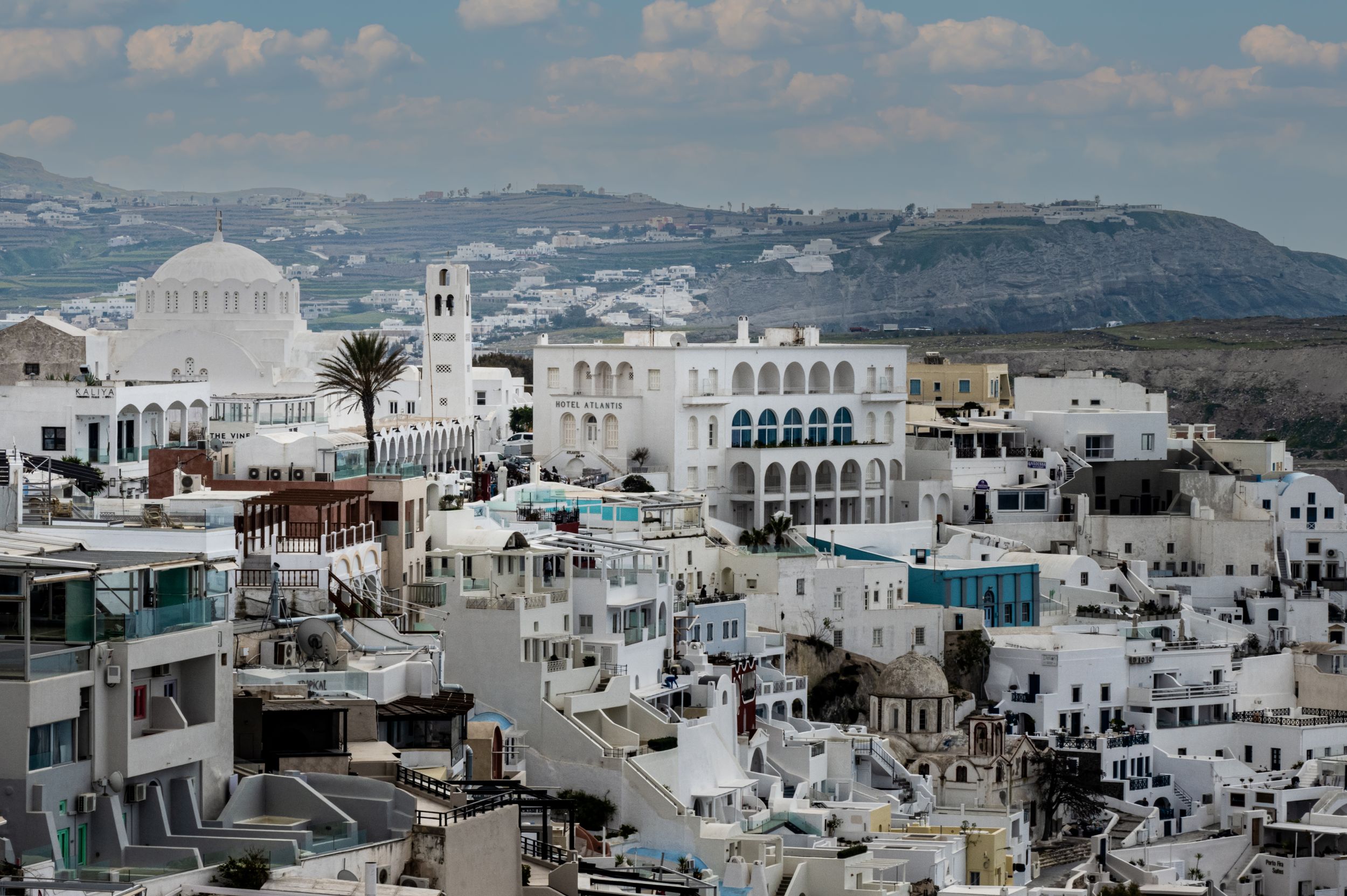 Panoramic view of white-washed buildings and churches in Mykonos, Greece with blue sky and mountains in background.