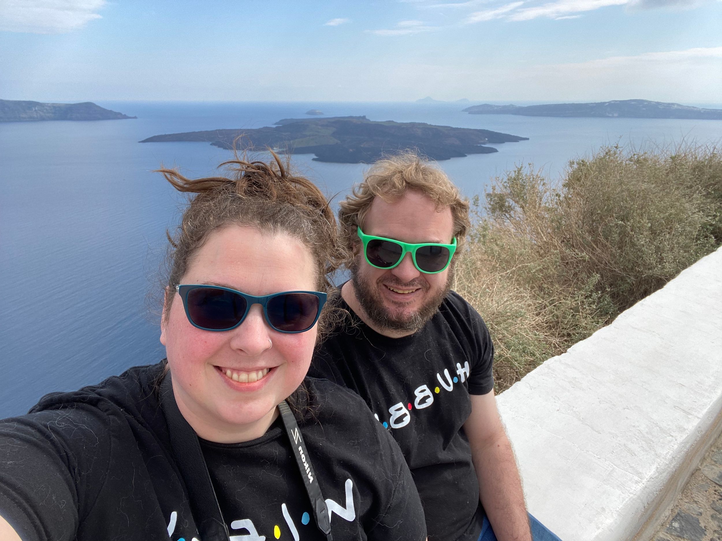 Two people in sunglasses take a selfie overlooking scenic Mediterranean islands and blue water on a sunny day.