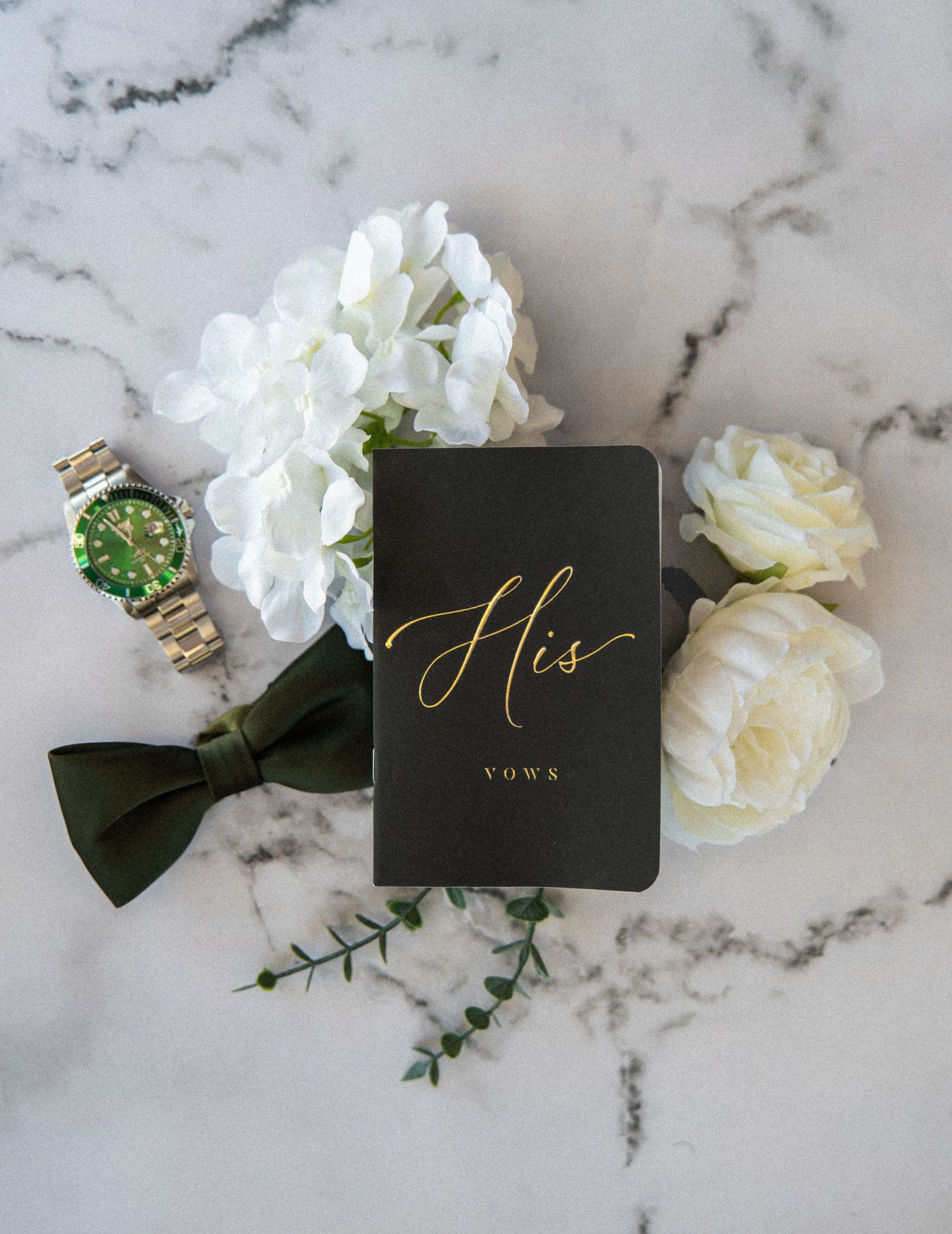 Black and gold His Vows book with white flowers, green bow tie, and Rolex watch on marble surface.