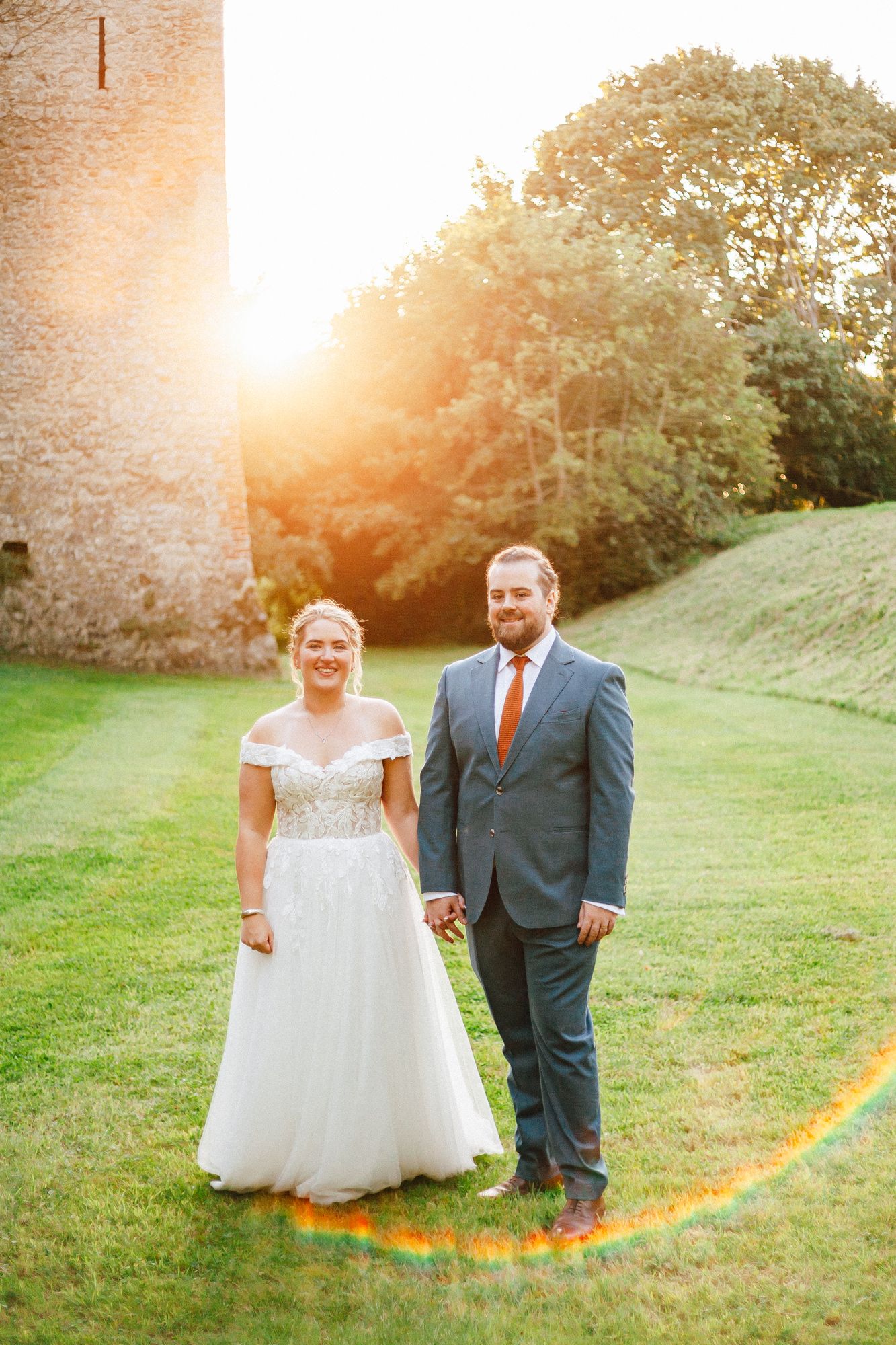 Bride and groom holding hands on green grass at golden hour near a stone castle wall.